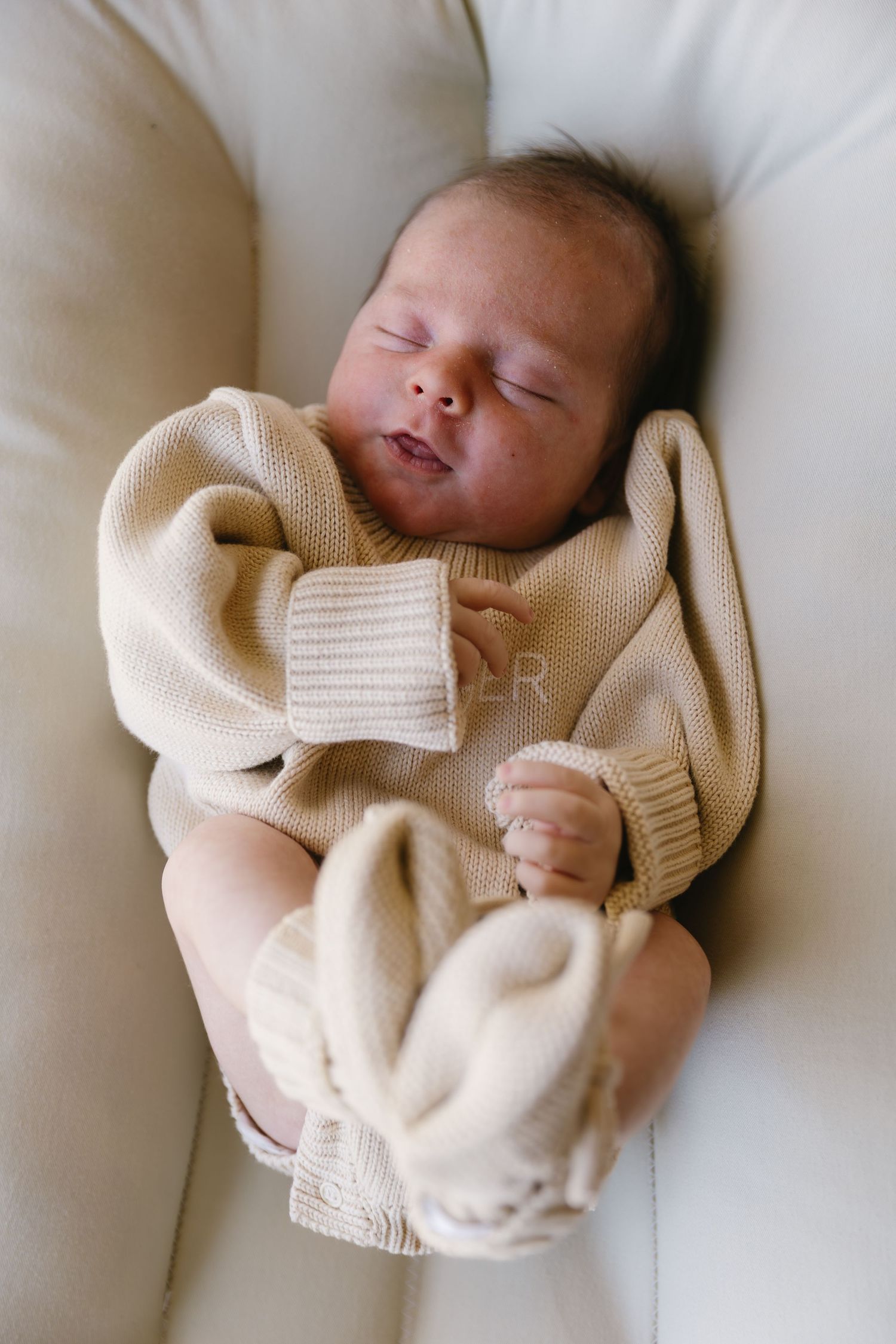 A sleeping newborn baby swaddled in a cozy cream-colored knit blanket lies peacefully on white bedding.
