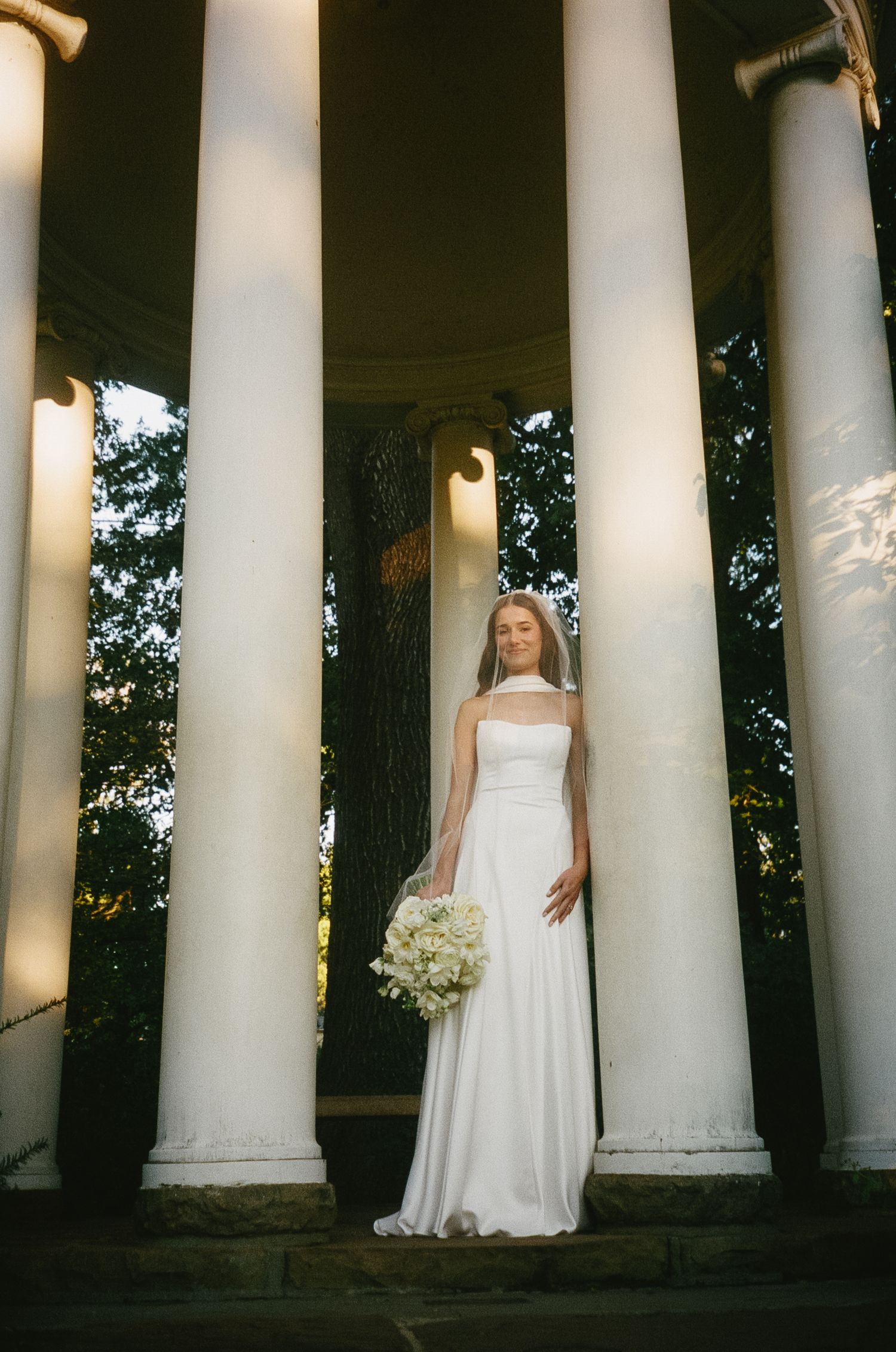 A bride in a white strapless gown holds a bouquet while standing between elegant white columns at dusk.