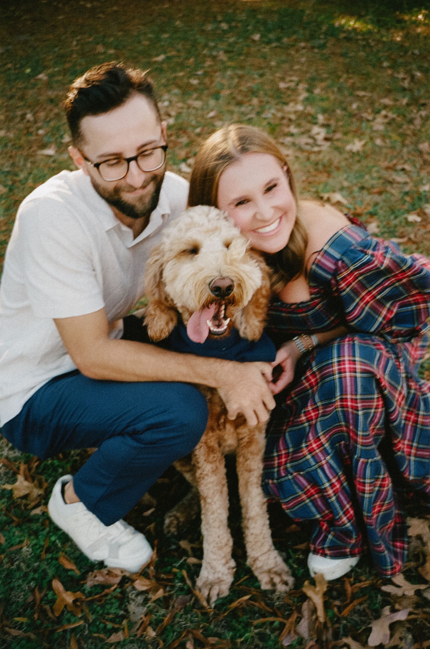 Couple sitting on grass with their goldendoodle dog in autumn setting wearing plaid and casual attire.