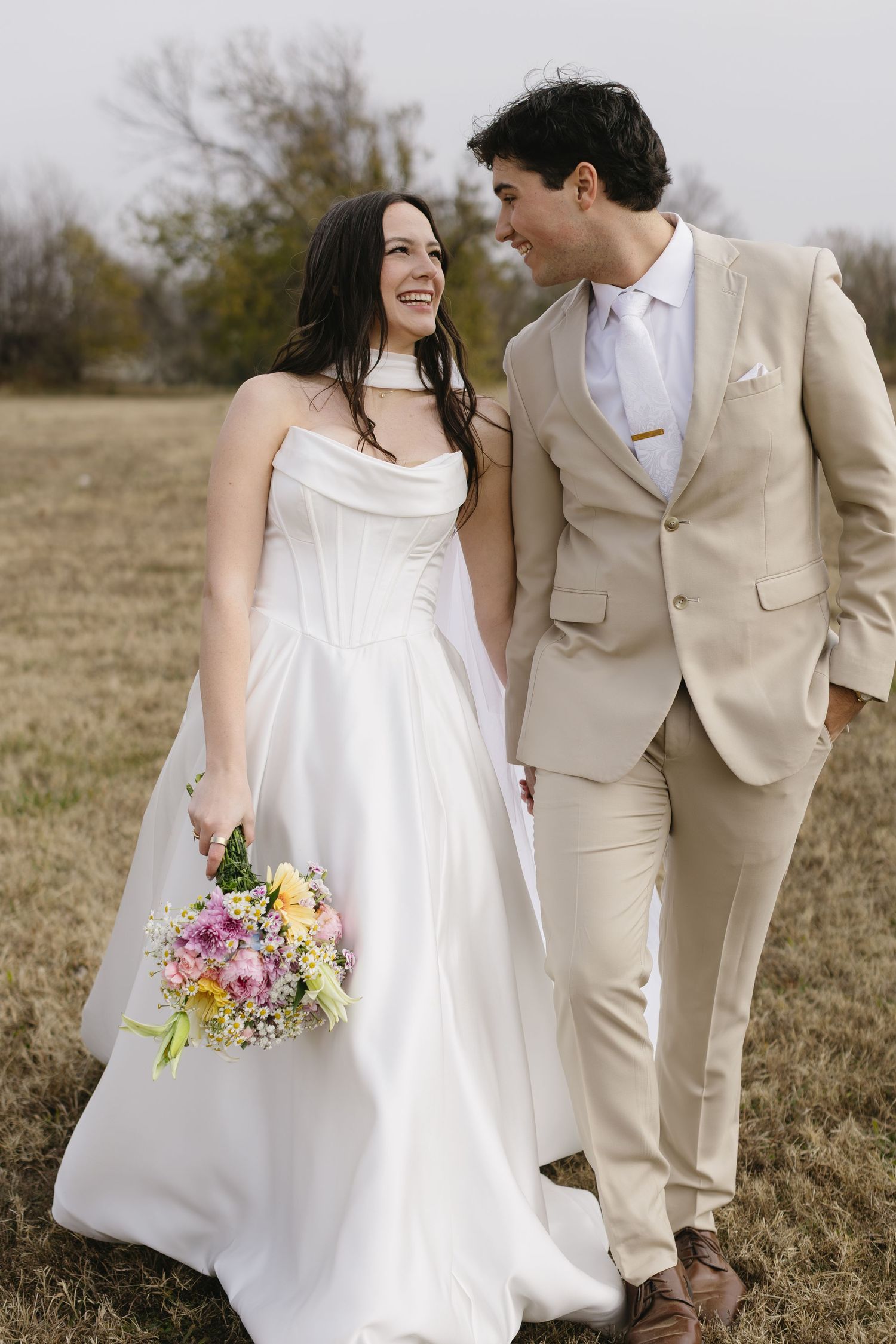 A couple in wedding attire shares a joyful moment in a rustic outdoor setting with a colorful bouquet.