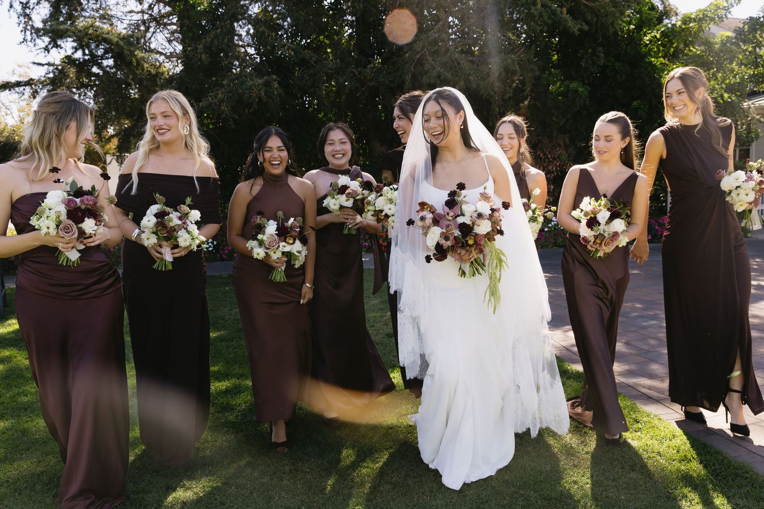 Wedding party in brown dresses and white gowns poses together outdoors on sunny day.