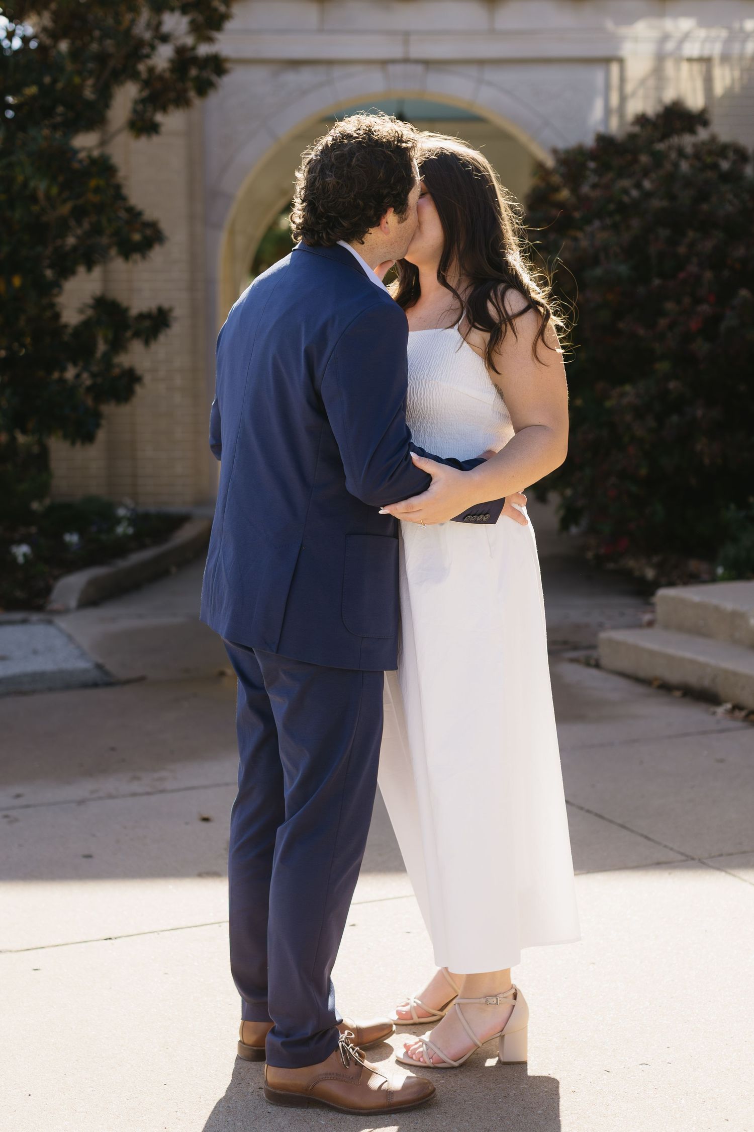 A couple embraces in formal wedding attire against a sunlit architectural backdrop.