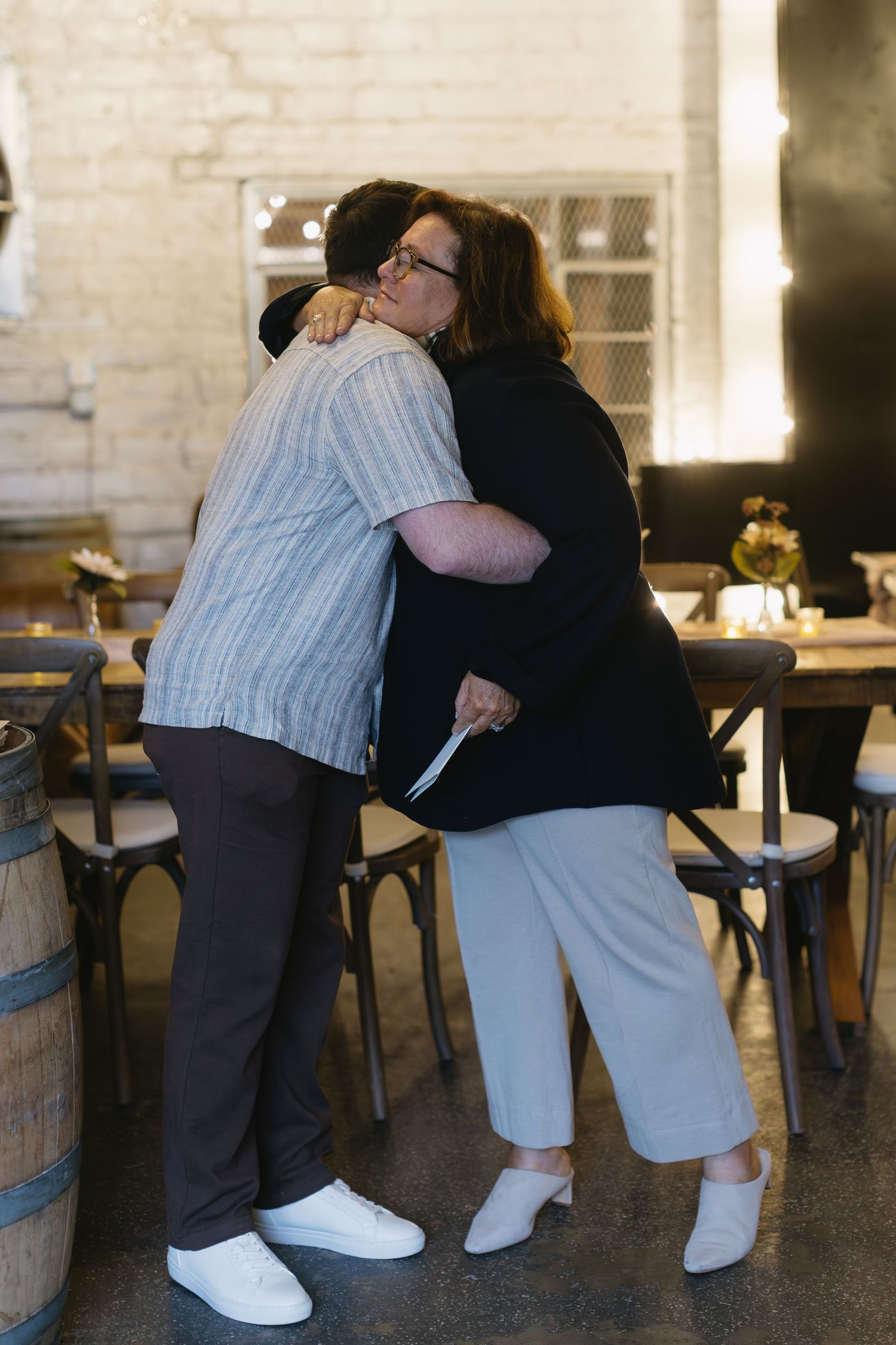 Two people sharing an emotional embrace in a rustic indoor setting with wooden barrels and chairs.