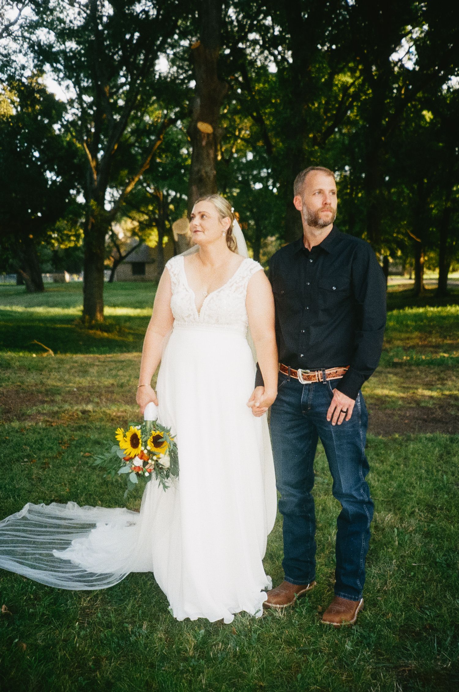 Wedding couple stands in a sunlit park with green trees and sunflowers in casual rustic attire.