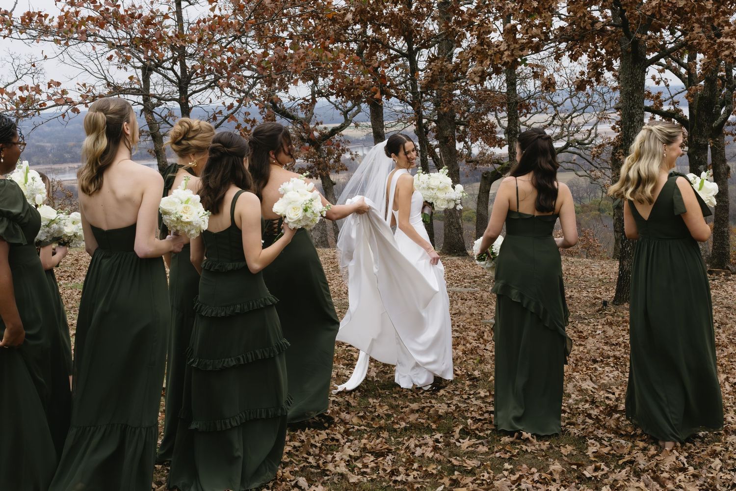 A wedding party poses in a line wearing black and white formal attire among autumn leaves on a hillside.