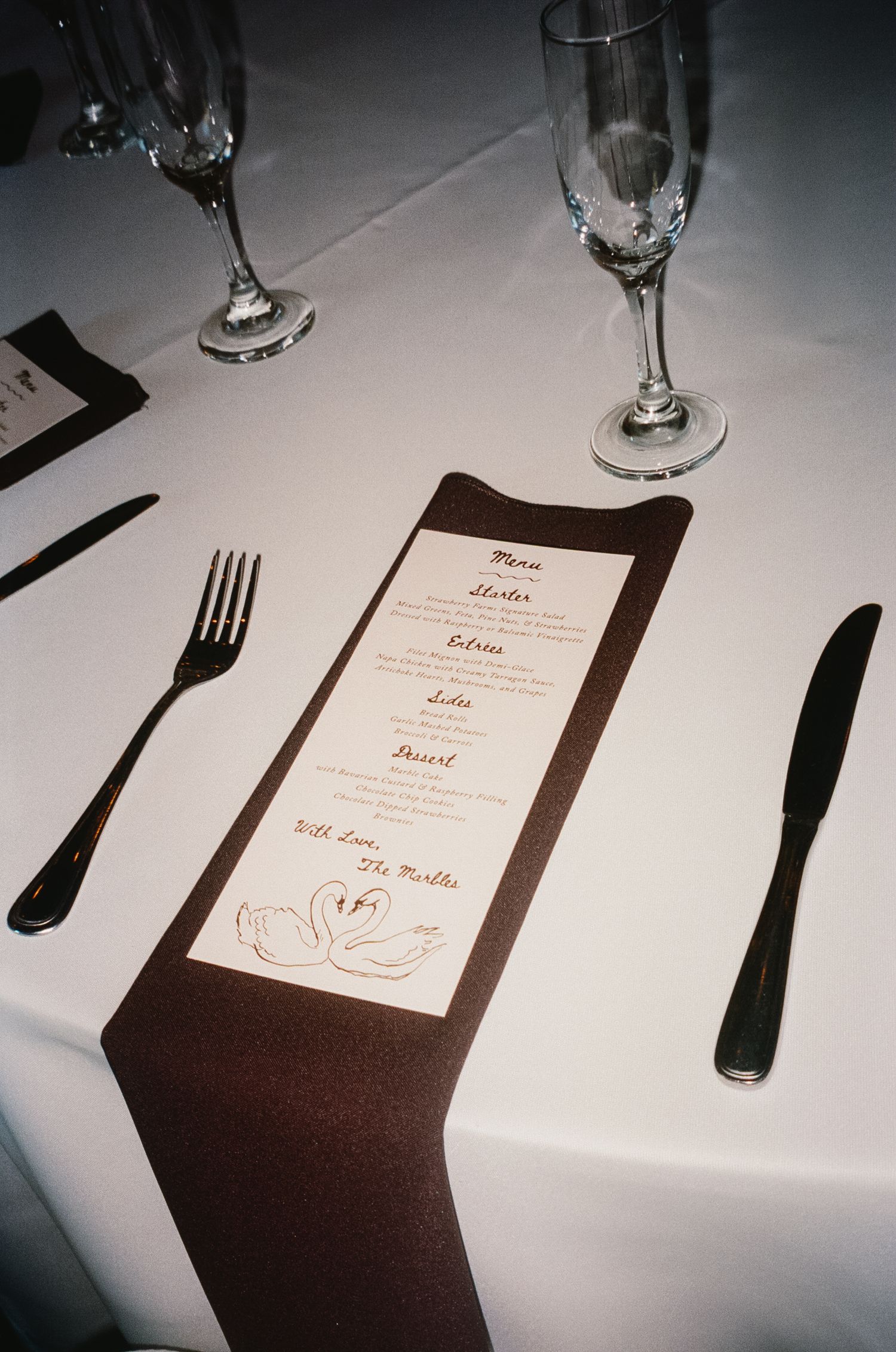 A formal place setting with menu card, silverware and wine glasses on a white tablecloth.