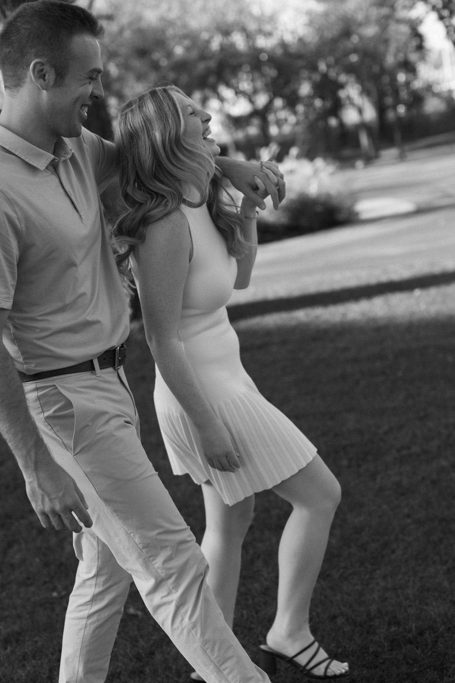 A couple in white outfits dances playfully together on a grassy lawn in black and white photography.