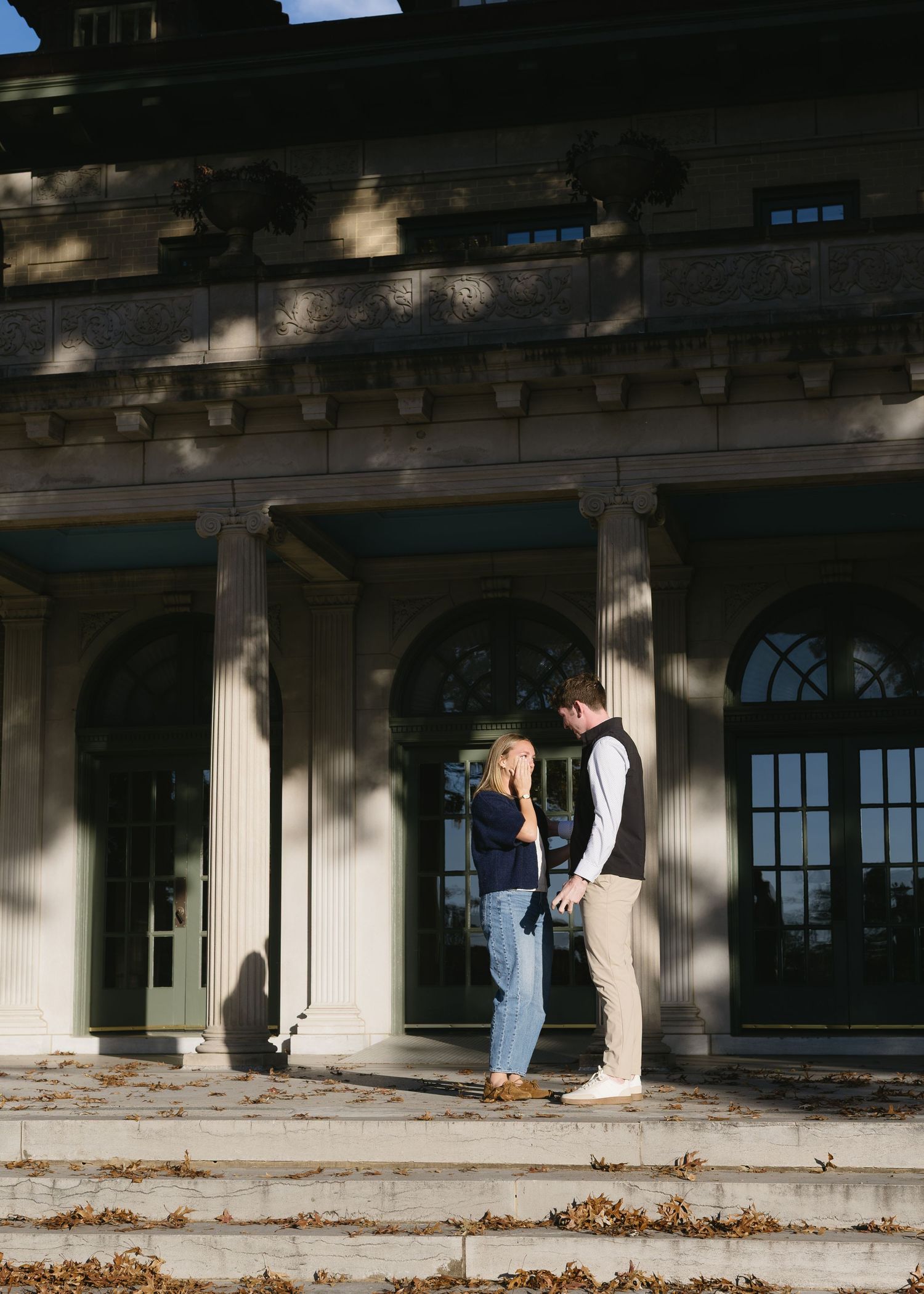 A couple embraces while standing on a porch with white columns and fallen leaves.