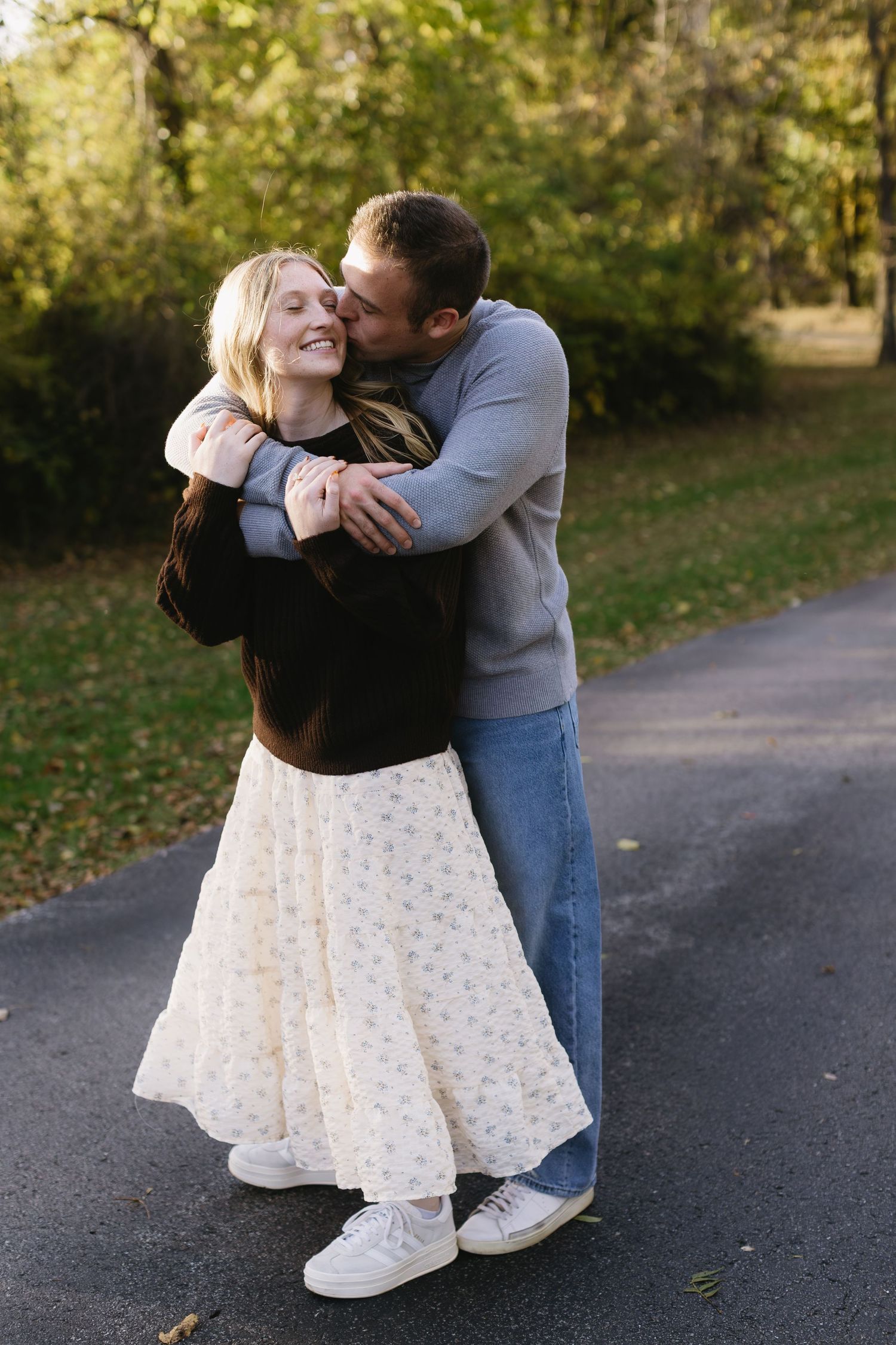 A couple dancing playfully together on a paved pathway surrounded by autumn trees.
