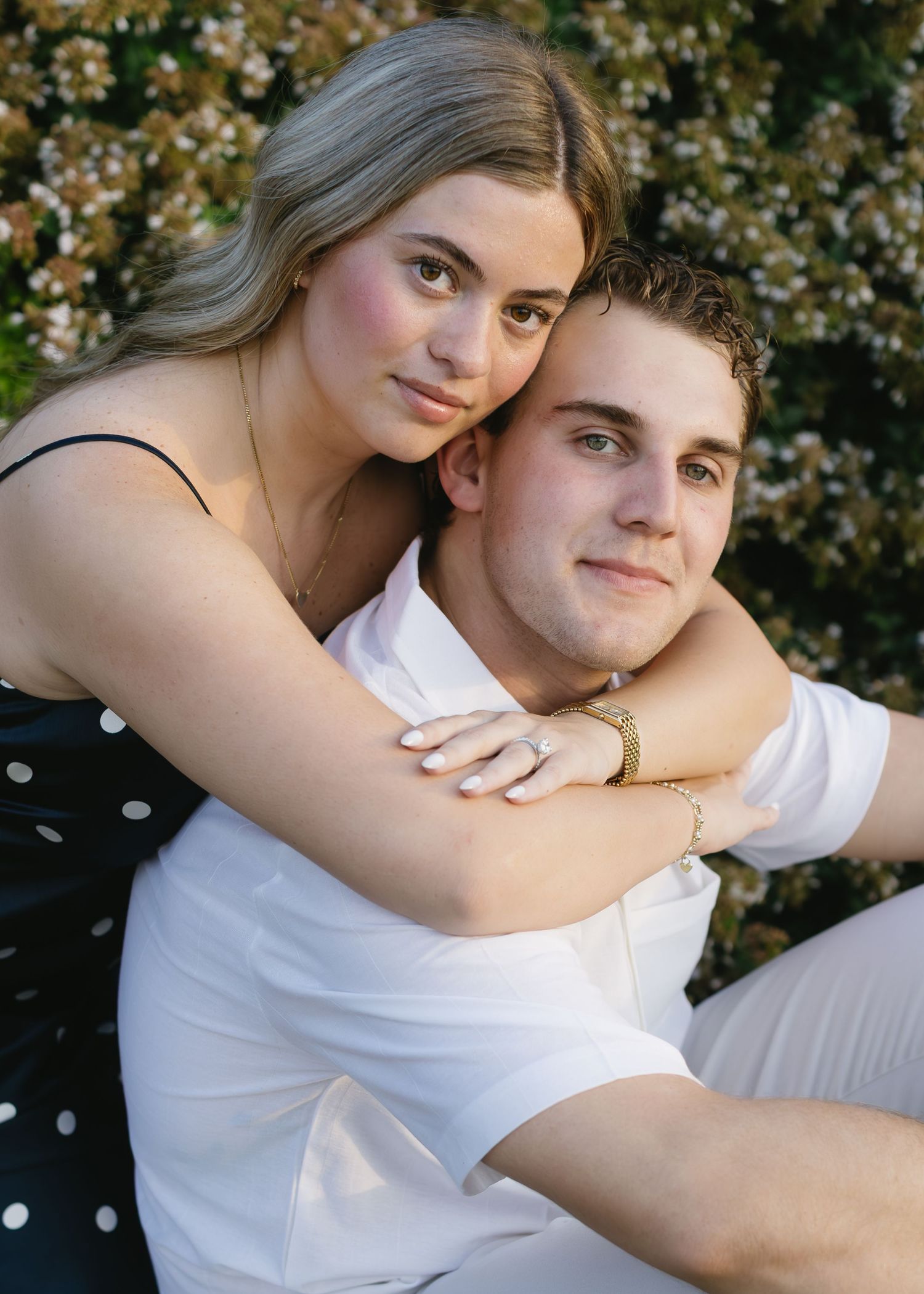 A couple embraces in a romantic outdoor portrait with blooming flowers in the background.