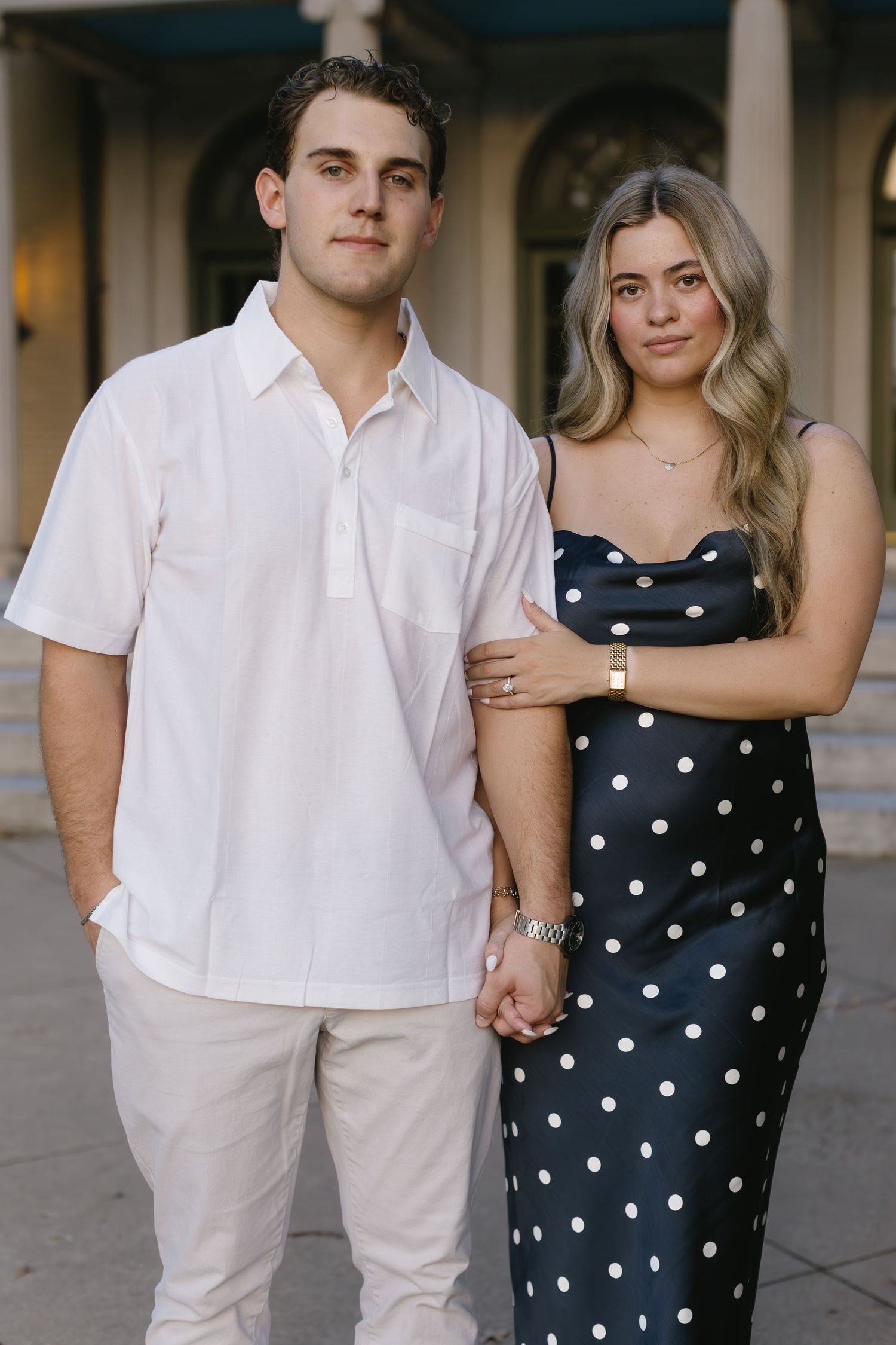 A couple poses together in coordinated outfits of white and navy polka dots against a columned backdrop.