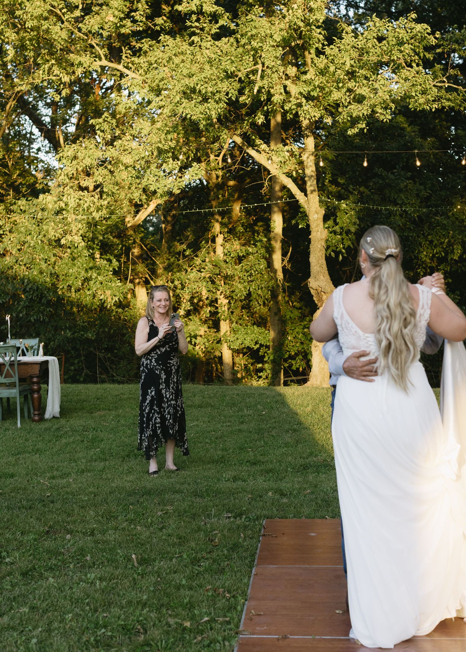 Wedding guests watching a couple dance on a wooden platform in a sunlit garden setting.