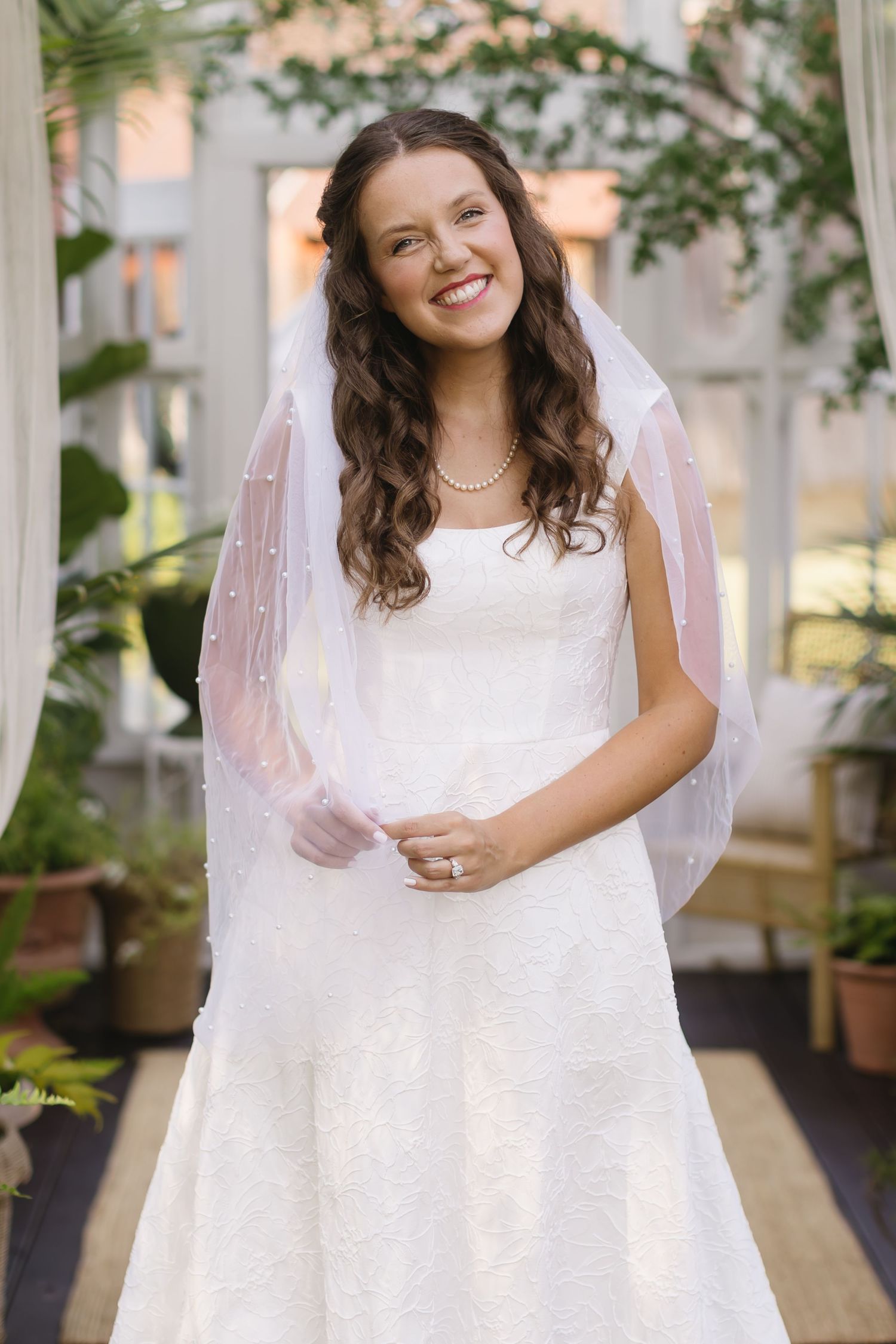 A bride wearing a classic white wedding gown with delicate pearl veil stands in a garden setting.