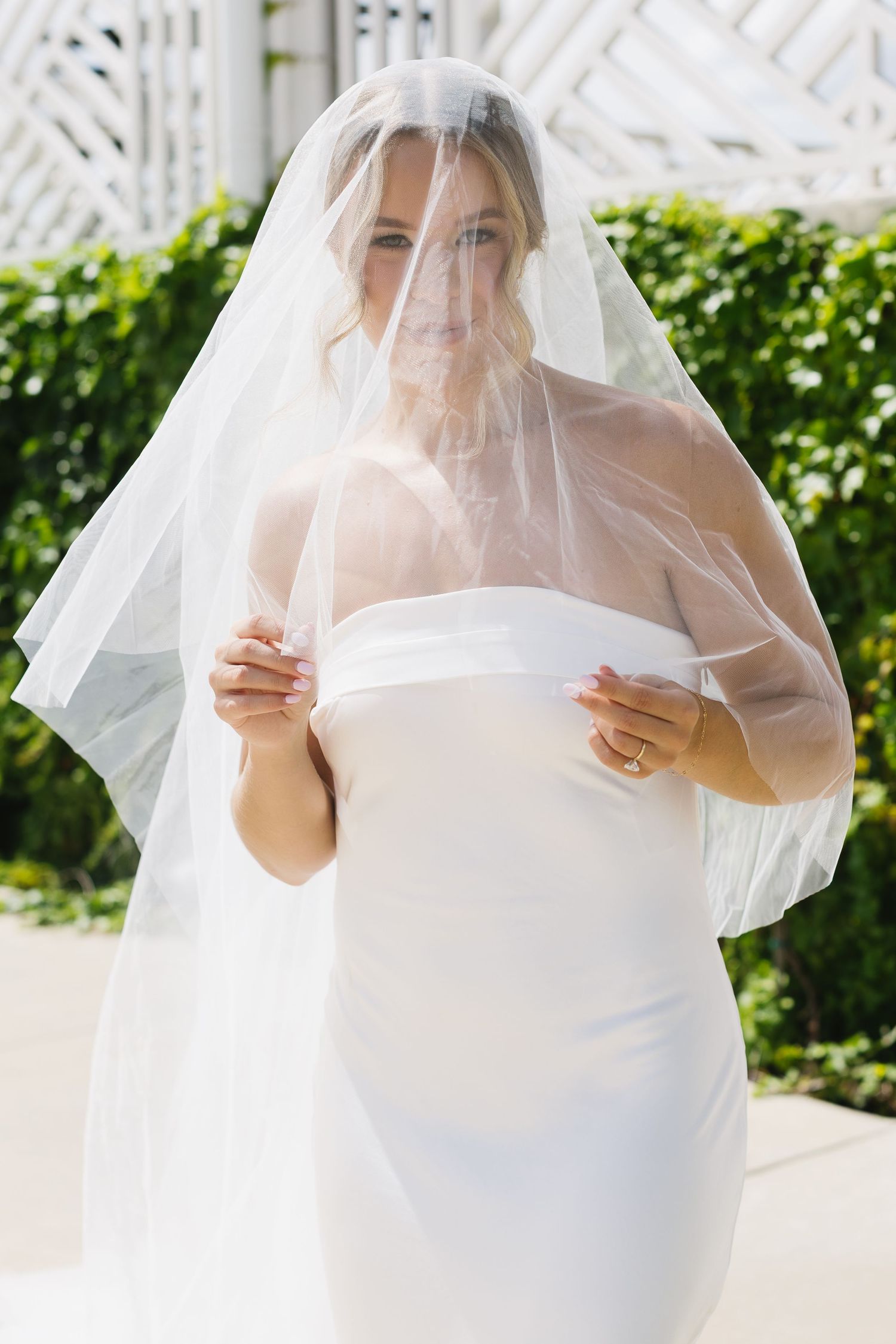 Bride in classic white dress plays with her veil outdoors on a sunny day.