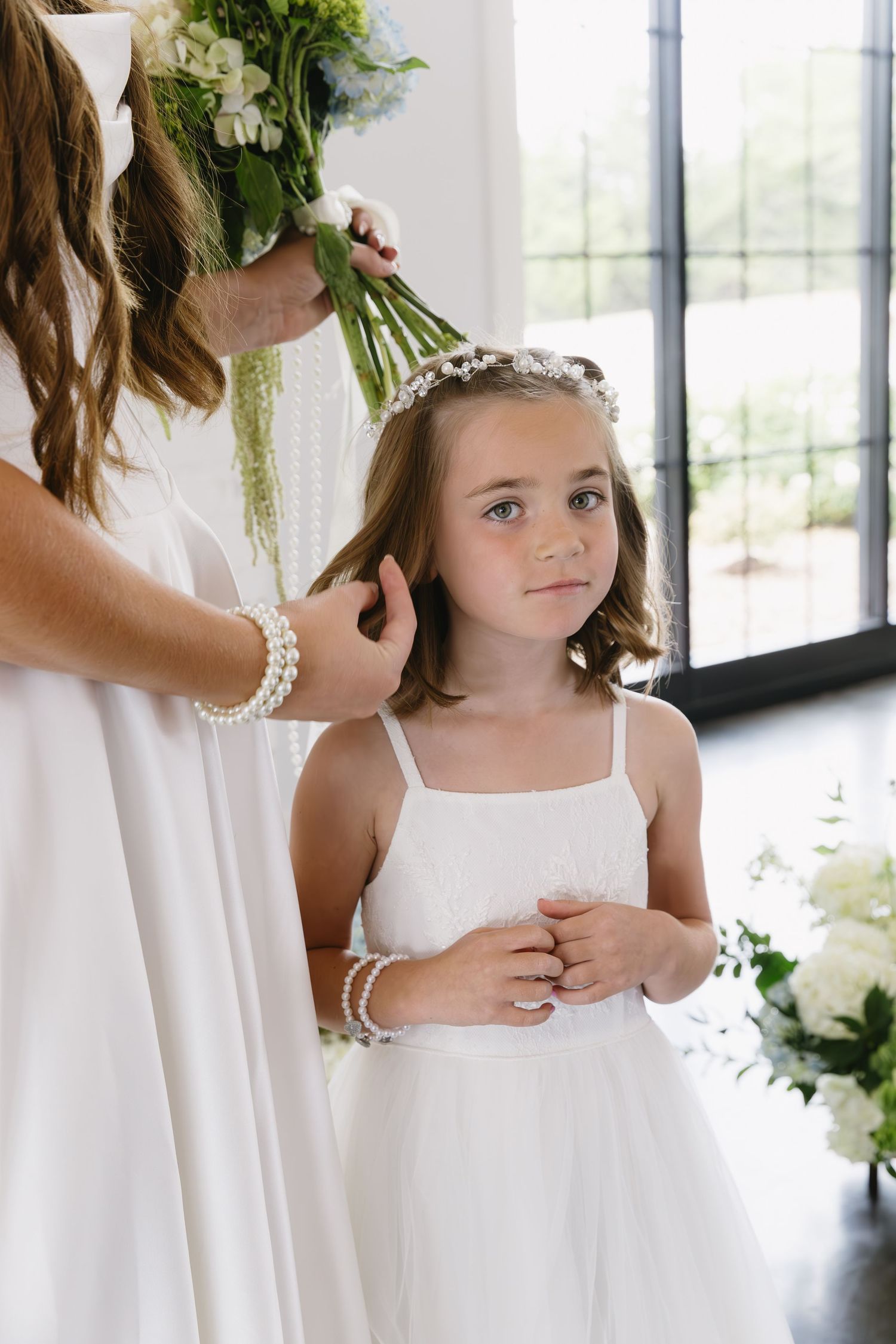 Young attendants in white dresses gather indoors near large windows during a formal event.