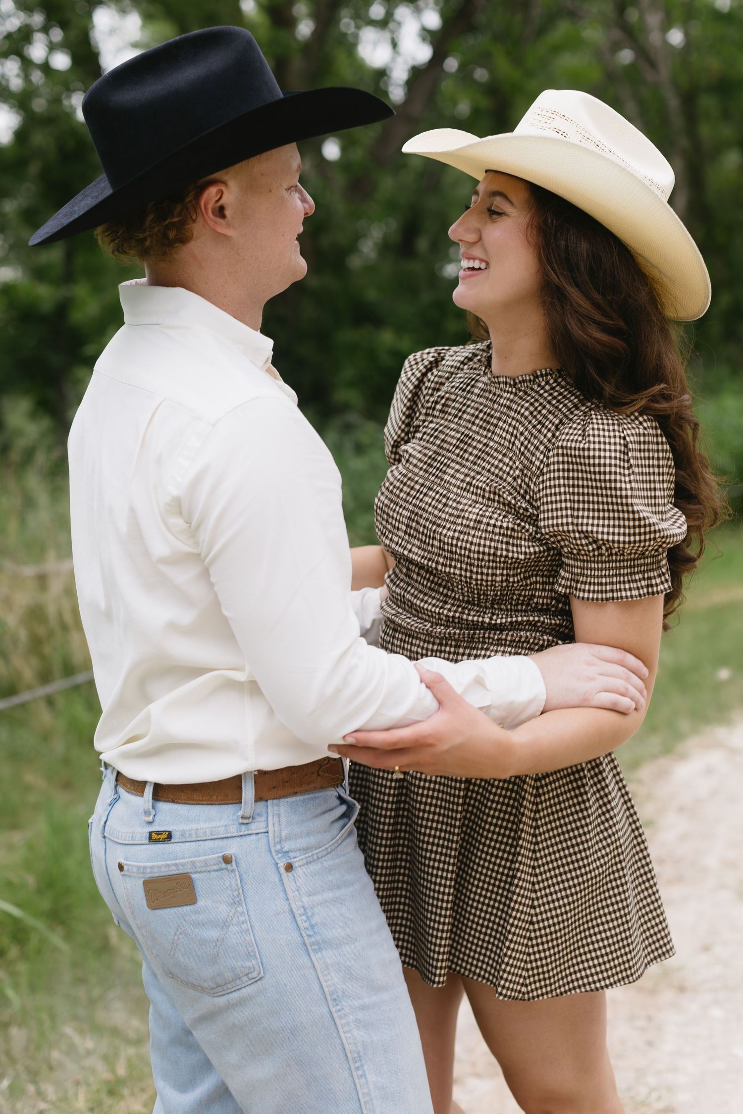 Romantic couple in cowboy hats and casual attire dance and laugh together on a dirt path.