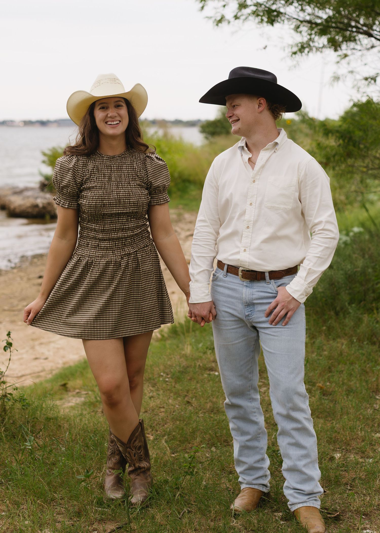 Couple in western wear strolls along beach shoreline wearing cowboy hats and boots.
