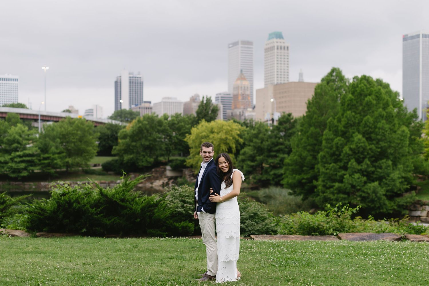 A romantic moment unfolds near a scenic pond with the city skyline visible through lush green trees in the background.