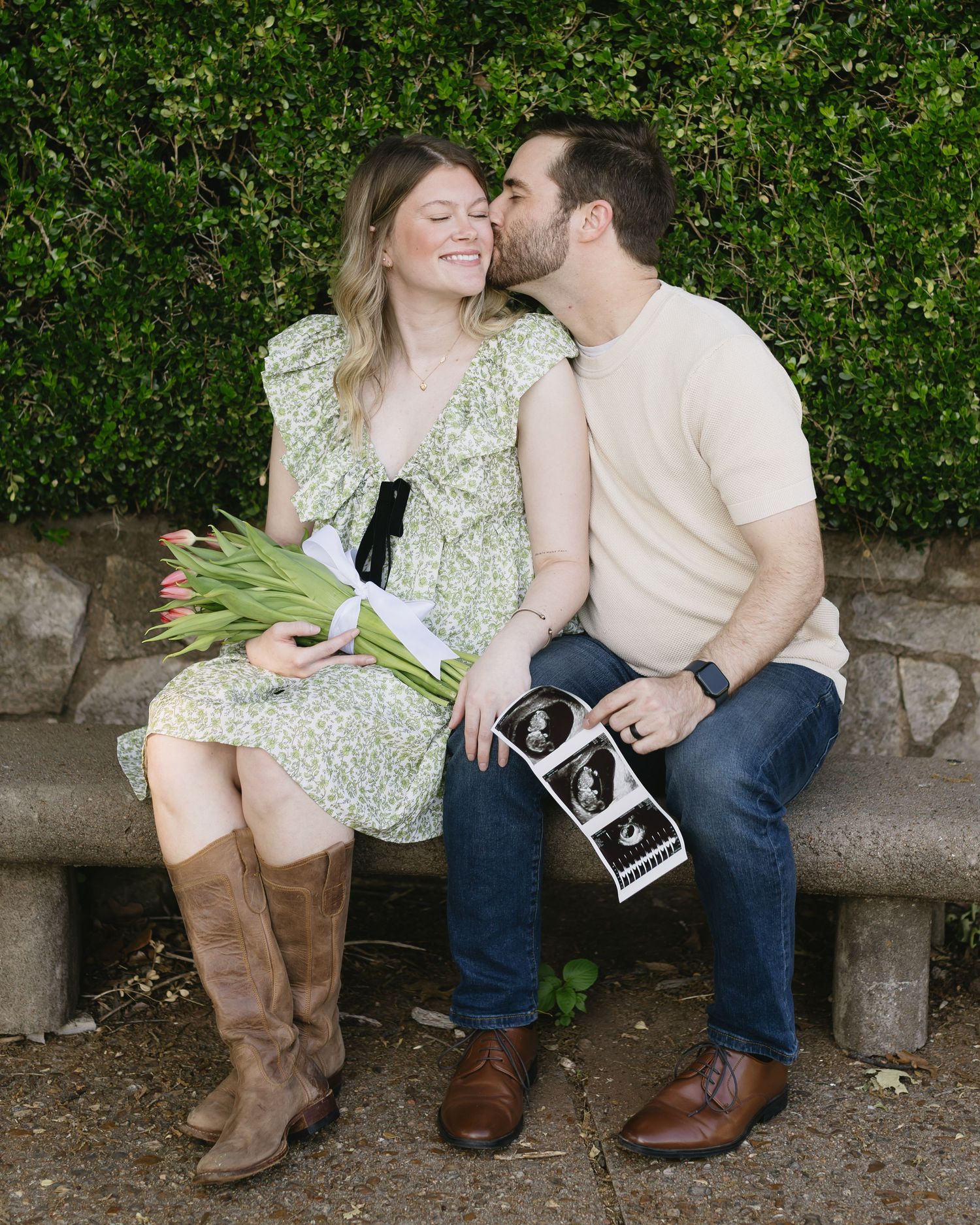 A couple sits casually on a stone wall surrounded by lush greenery while sharing a tender moment together.
