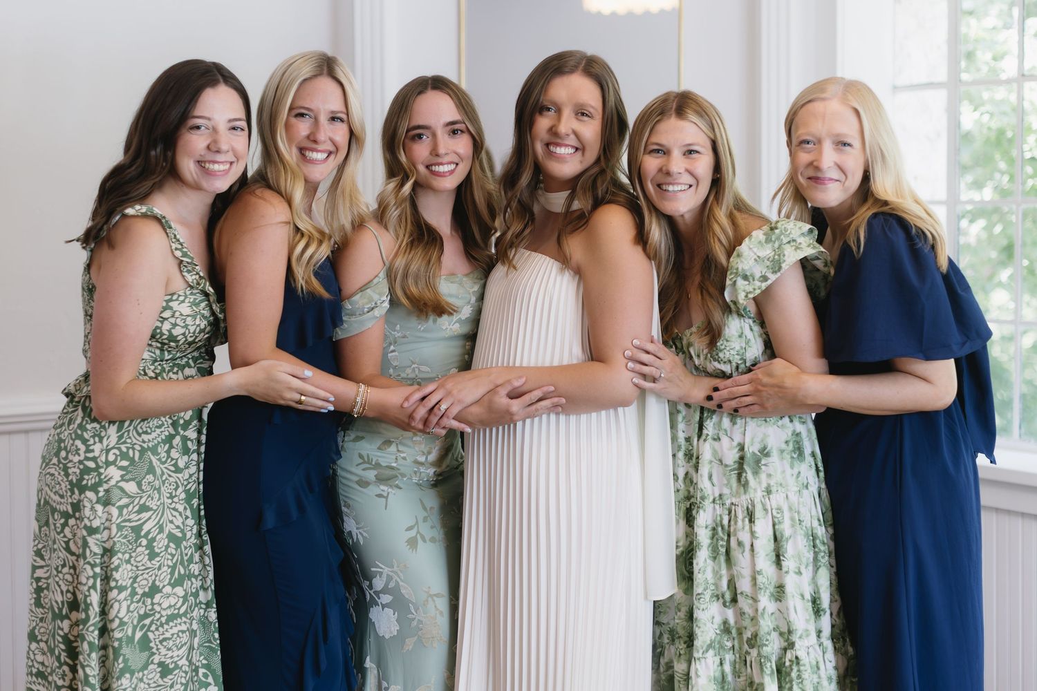 A wedding party poses in alternating navy blue and mint green floral dresses standing in front of white columns.