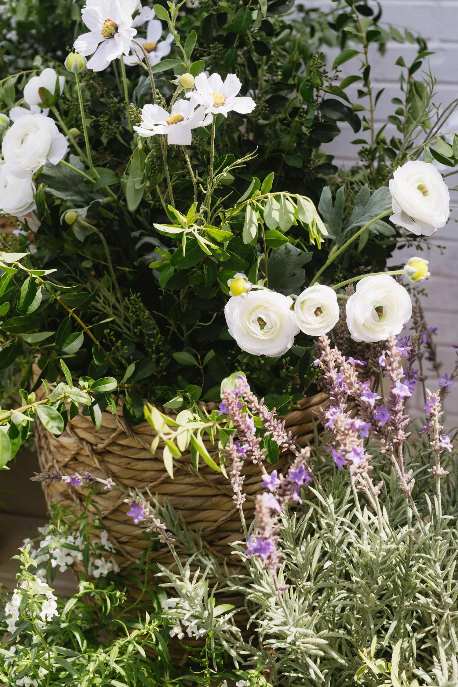 White flowers and purple lavender blooms arranged in a rustic woven basket against green foliage.