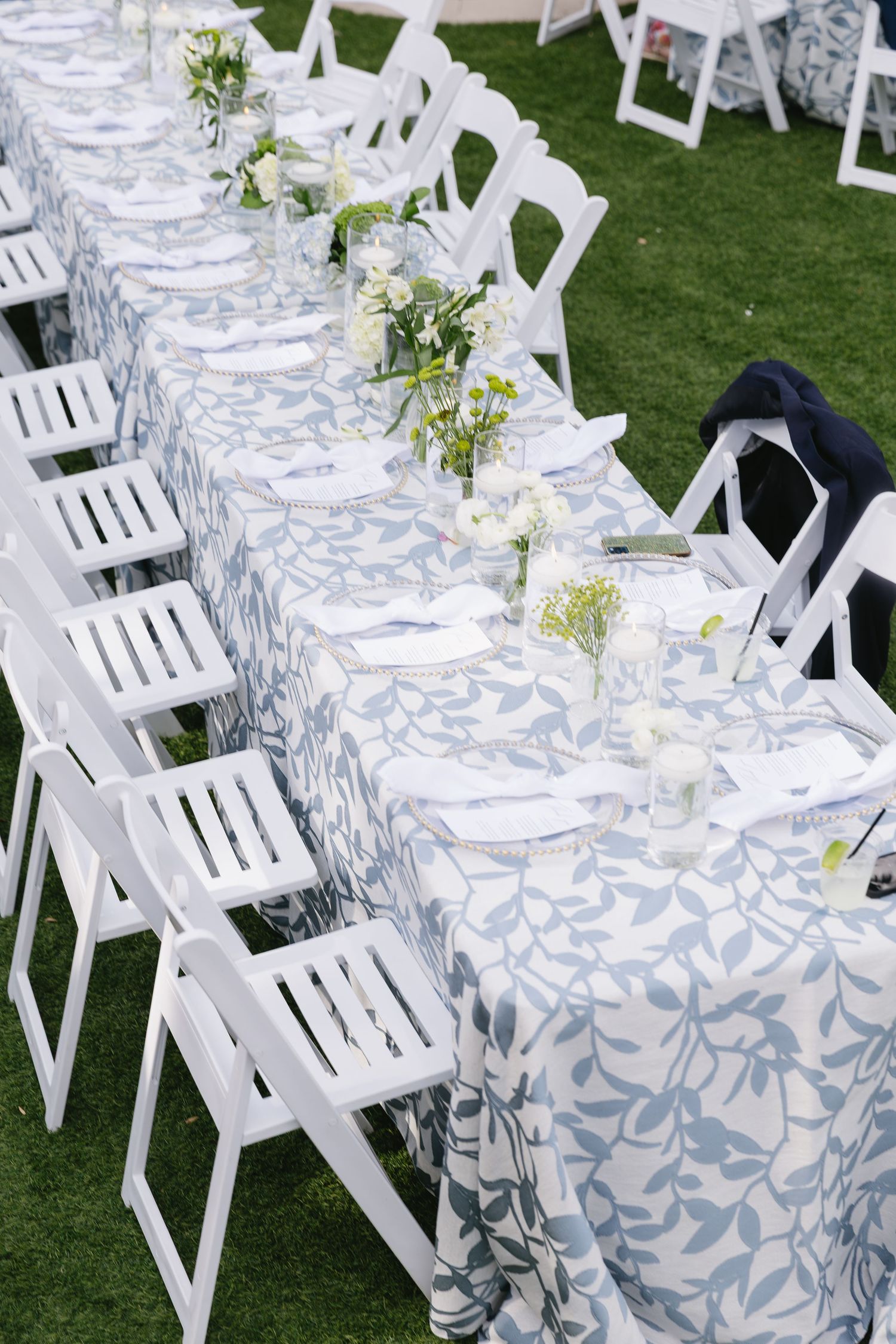 Outdoor wedding reception setup with white chairs and blue patterned tablecloths on grass.