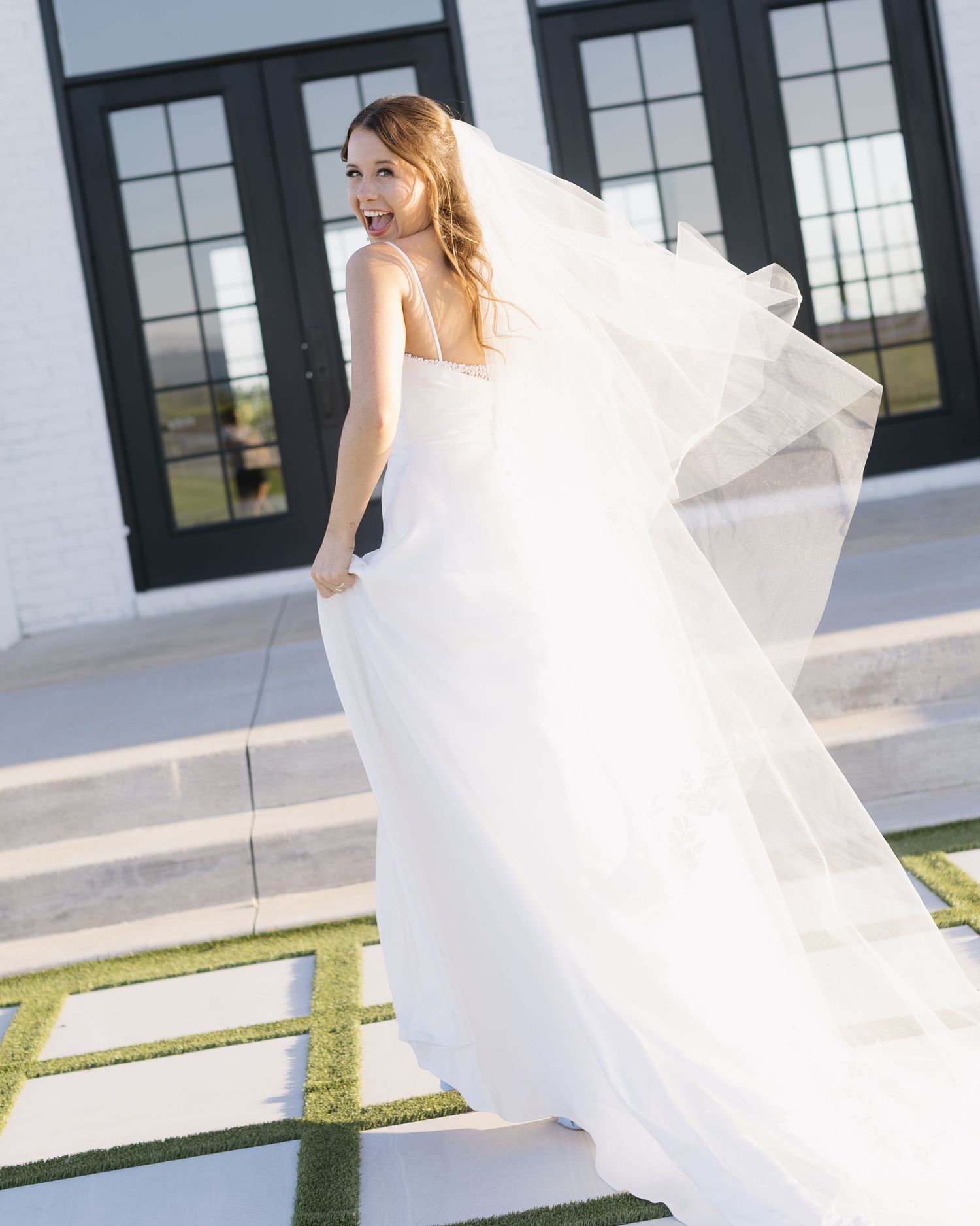 A bride in a flowing white gown twirls outside a modern white building with black windows.