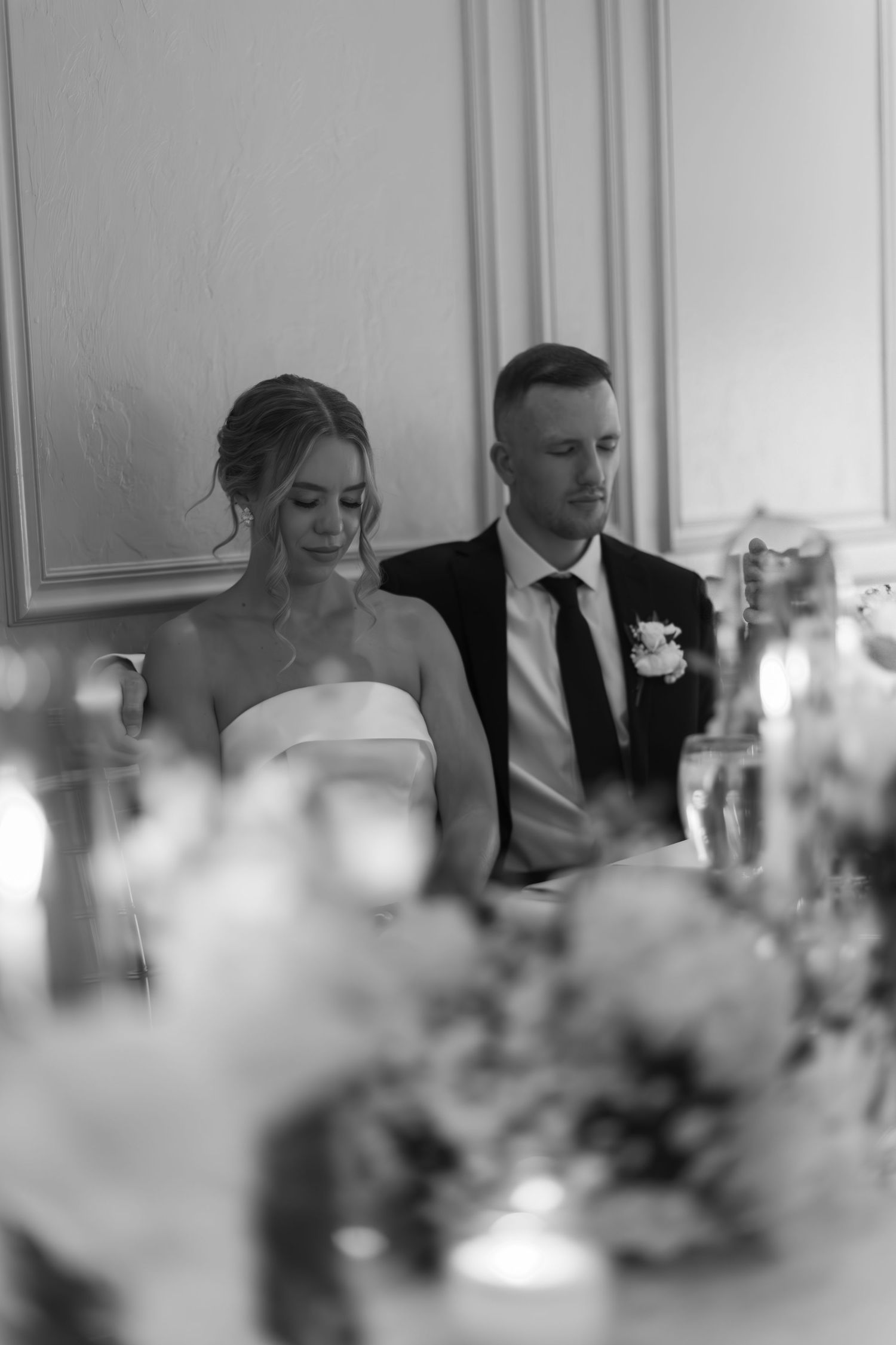 Black and white photo of a couple seated at an elegant wedding reception table with floral arrangements visible in foreground.