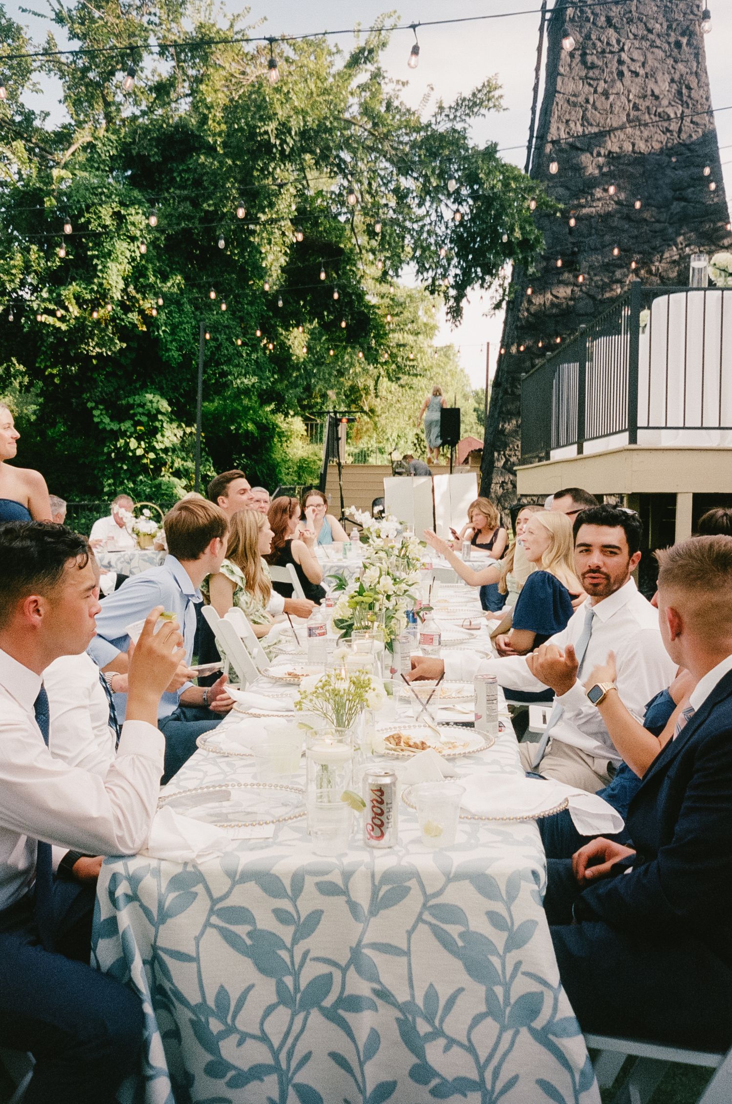 Guests seated at an elegant outdoor wedding reception table with floral centerpieces and a patterned tablecloth.