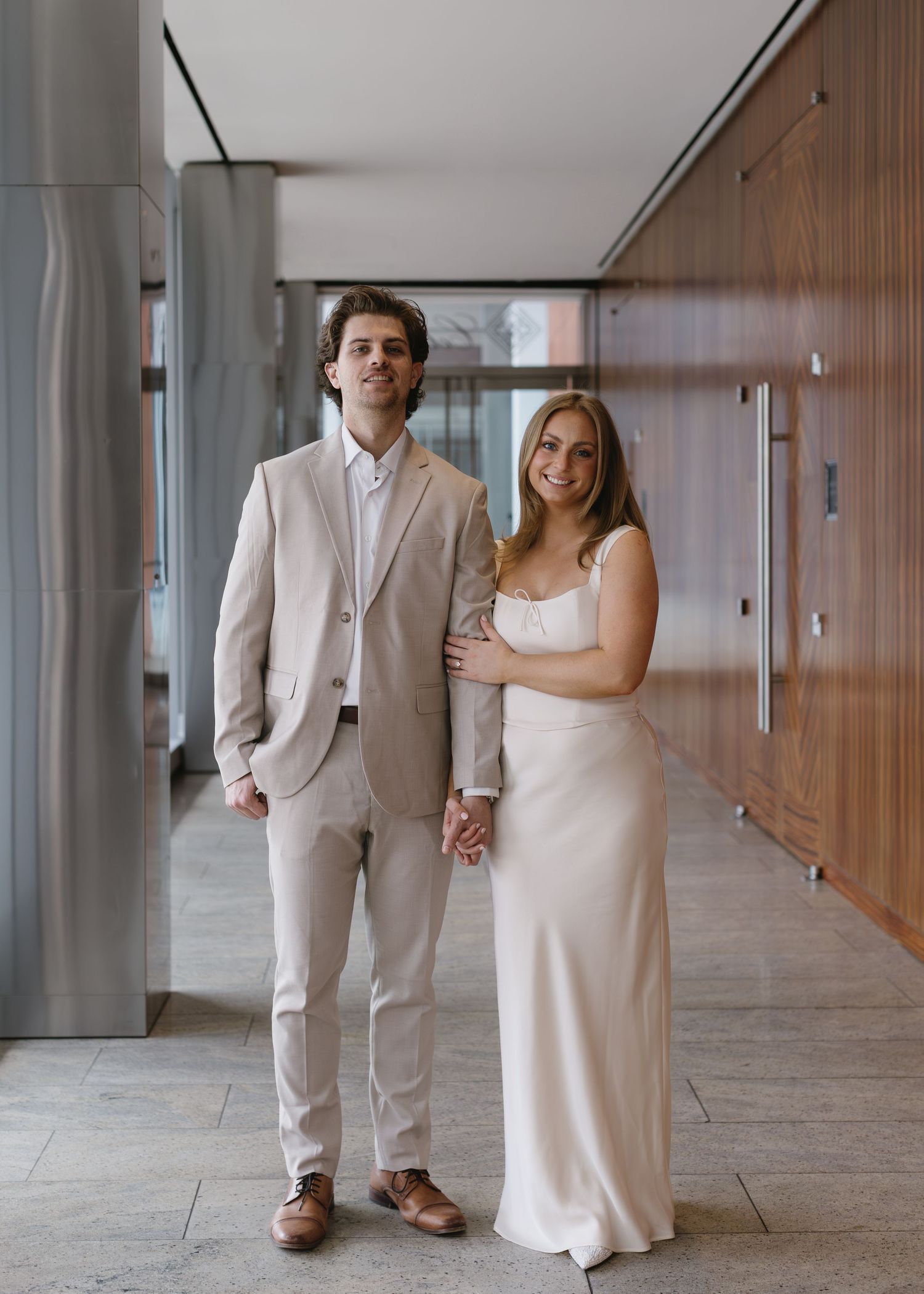 A couple in matching light colored formal wear pose together in an elegant hallway.