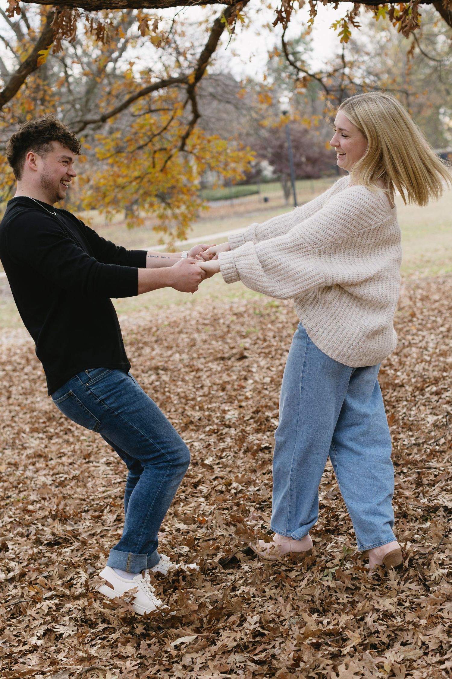 A playful couple in jeans and sweaters dance together in fallen autumn leaves.