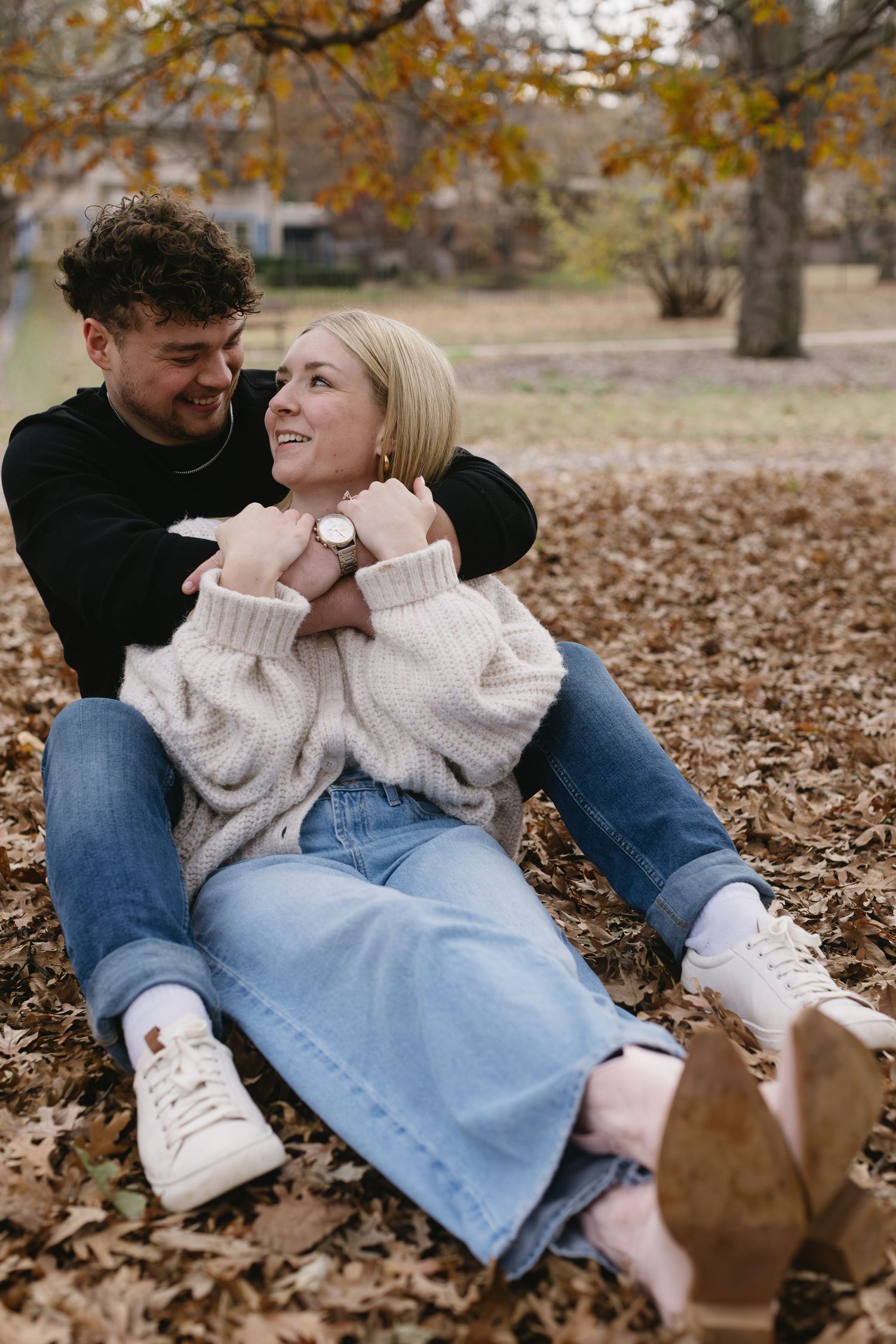 A couple sitting together on fallen autumn leaves wearing casual sweaters and jeans.