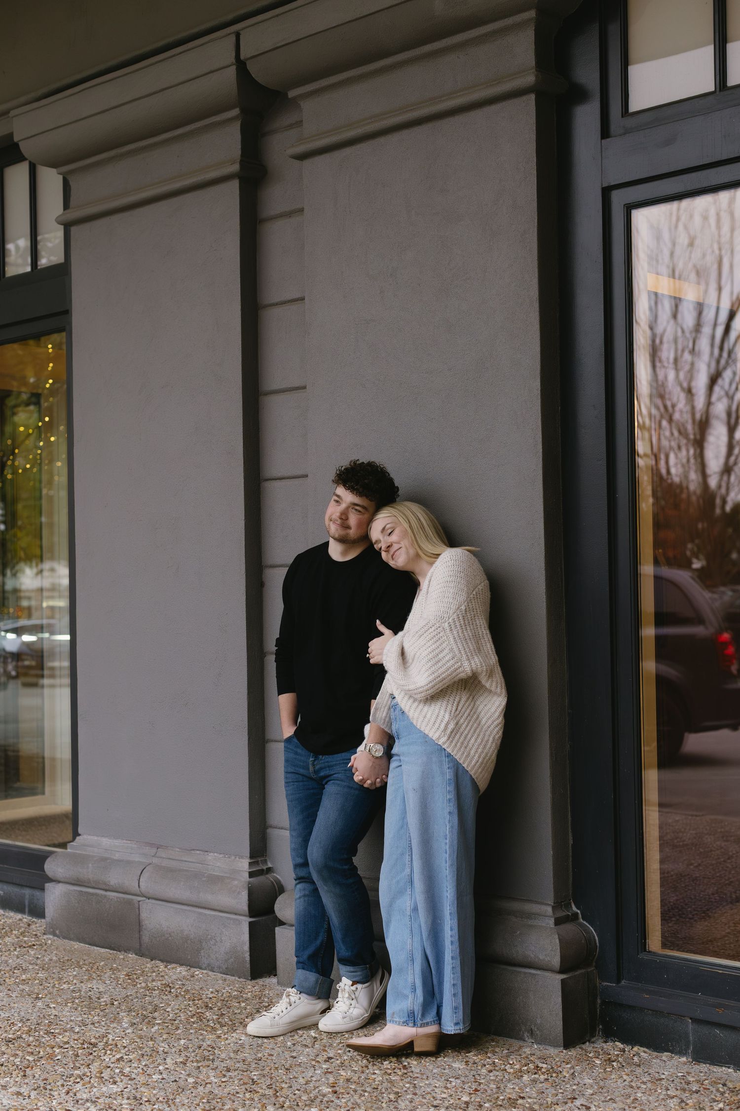 A couple poses together against a grey building wall in an urban setting.