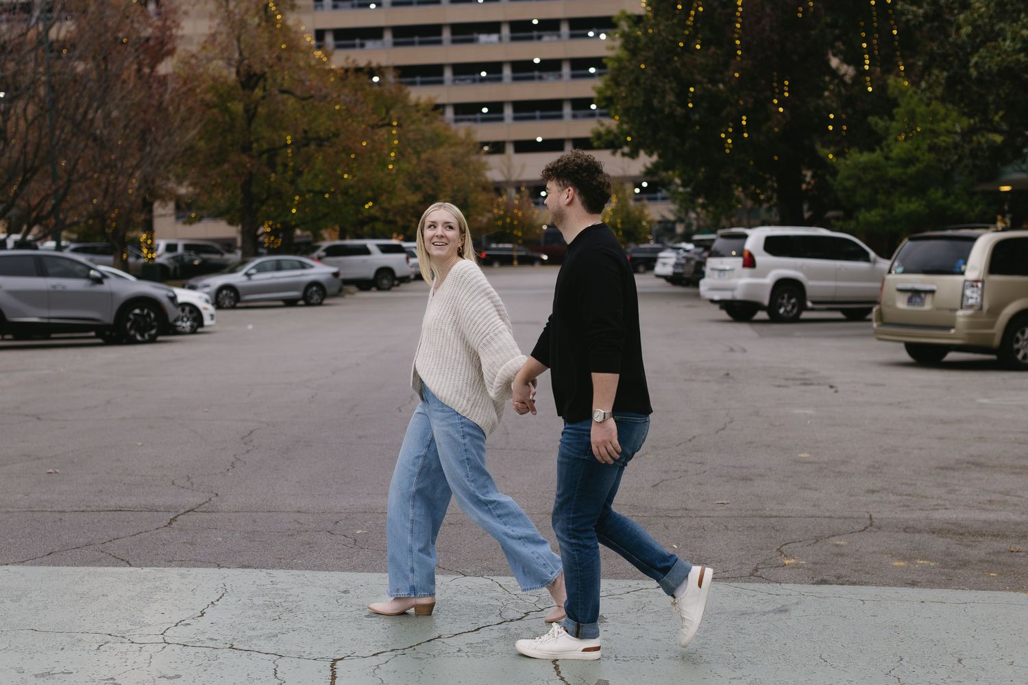People walking through a parking lot with tall buildings and autumn trees in the background.