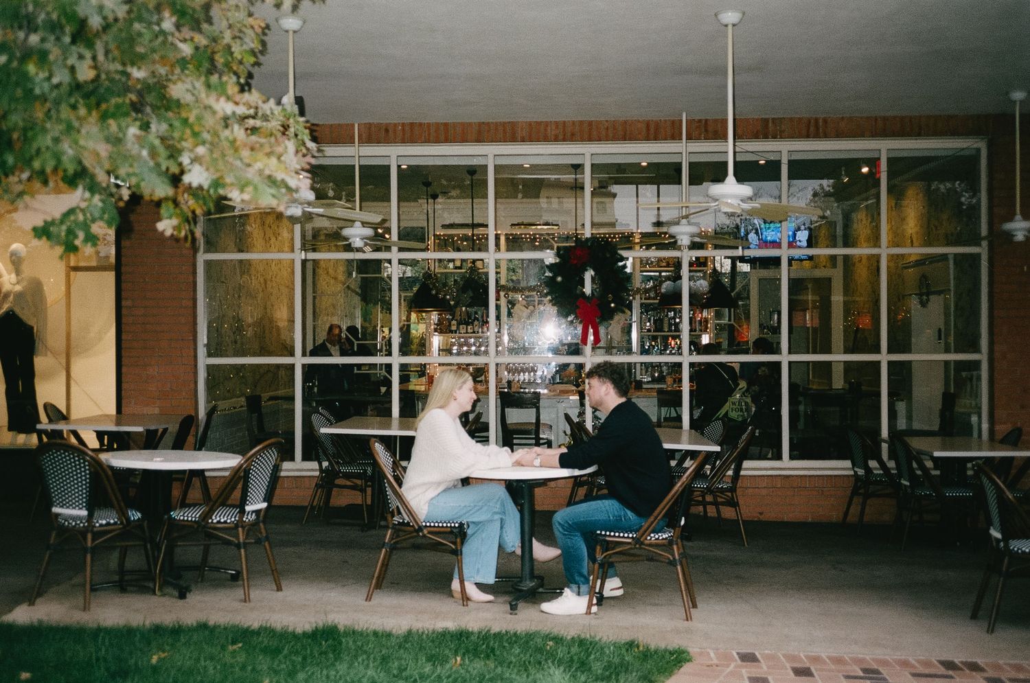 A couple sitting at an outdoor cafe table with festive lights visible in the background.