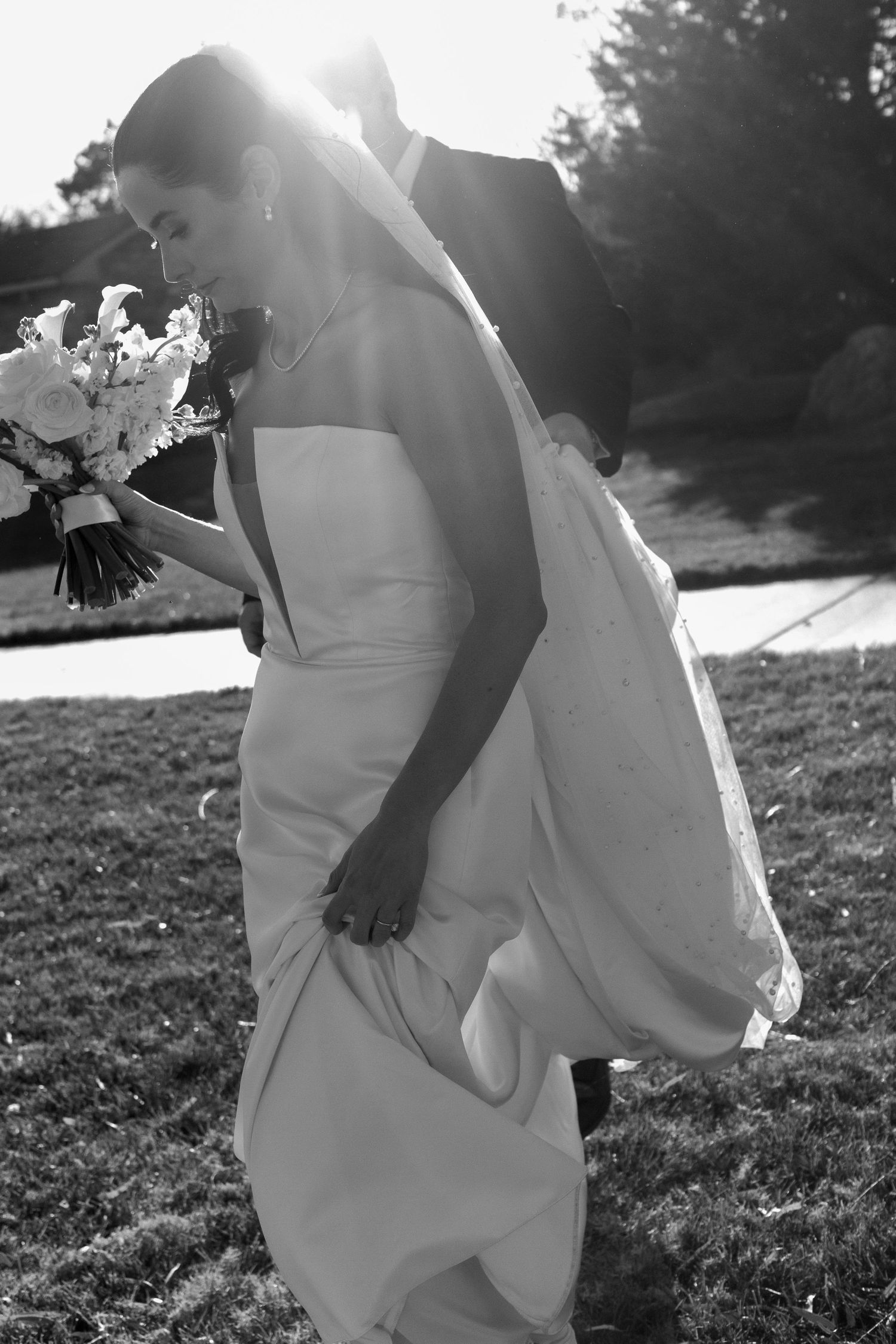 Black and white photo of newlyweds embracing in sunlit outdoor setting.