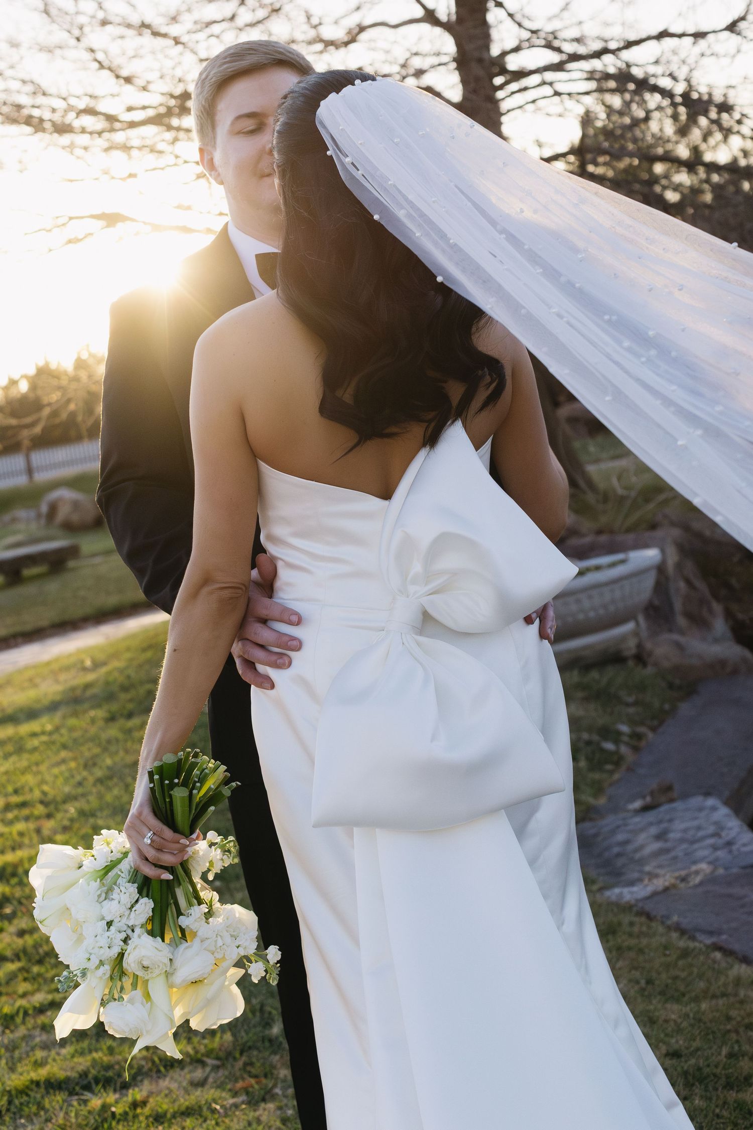 Elegant wedding couple poses in golden sunlight with bride holding white bouquet.