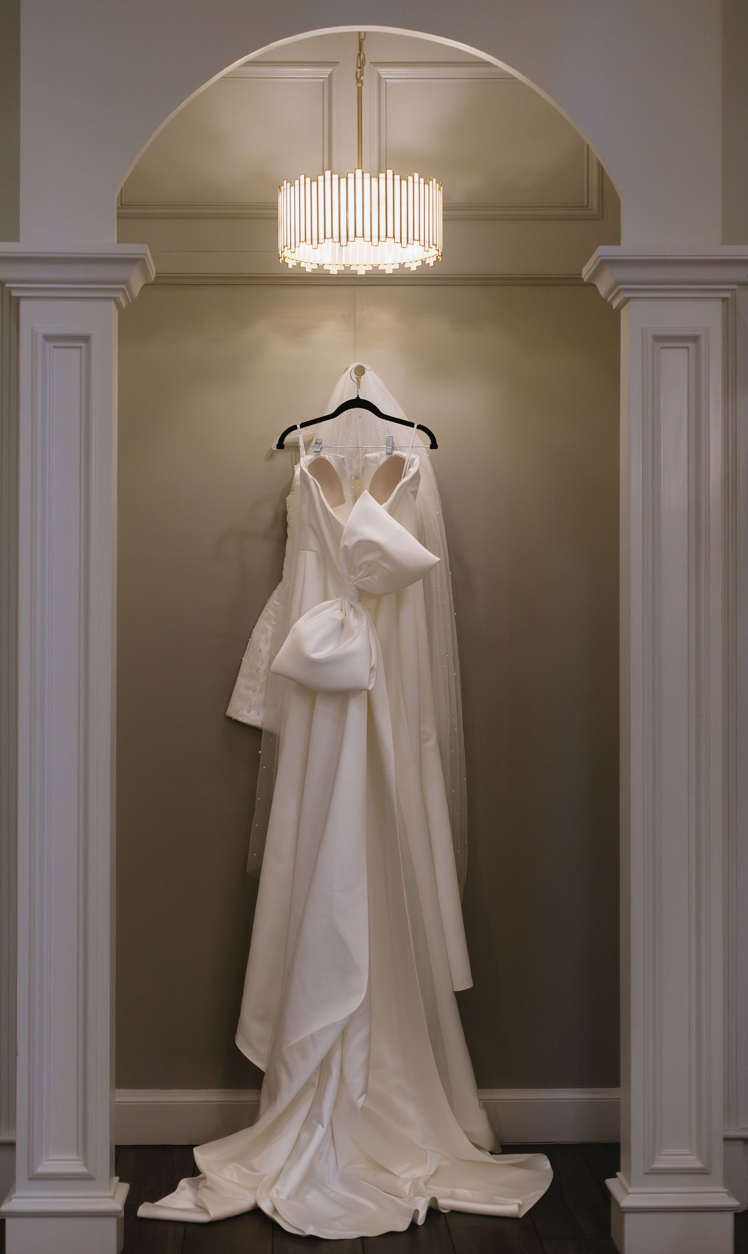 An elegant white wedding dress hanging in an arched alcove beneath a modern chandelier.