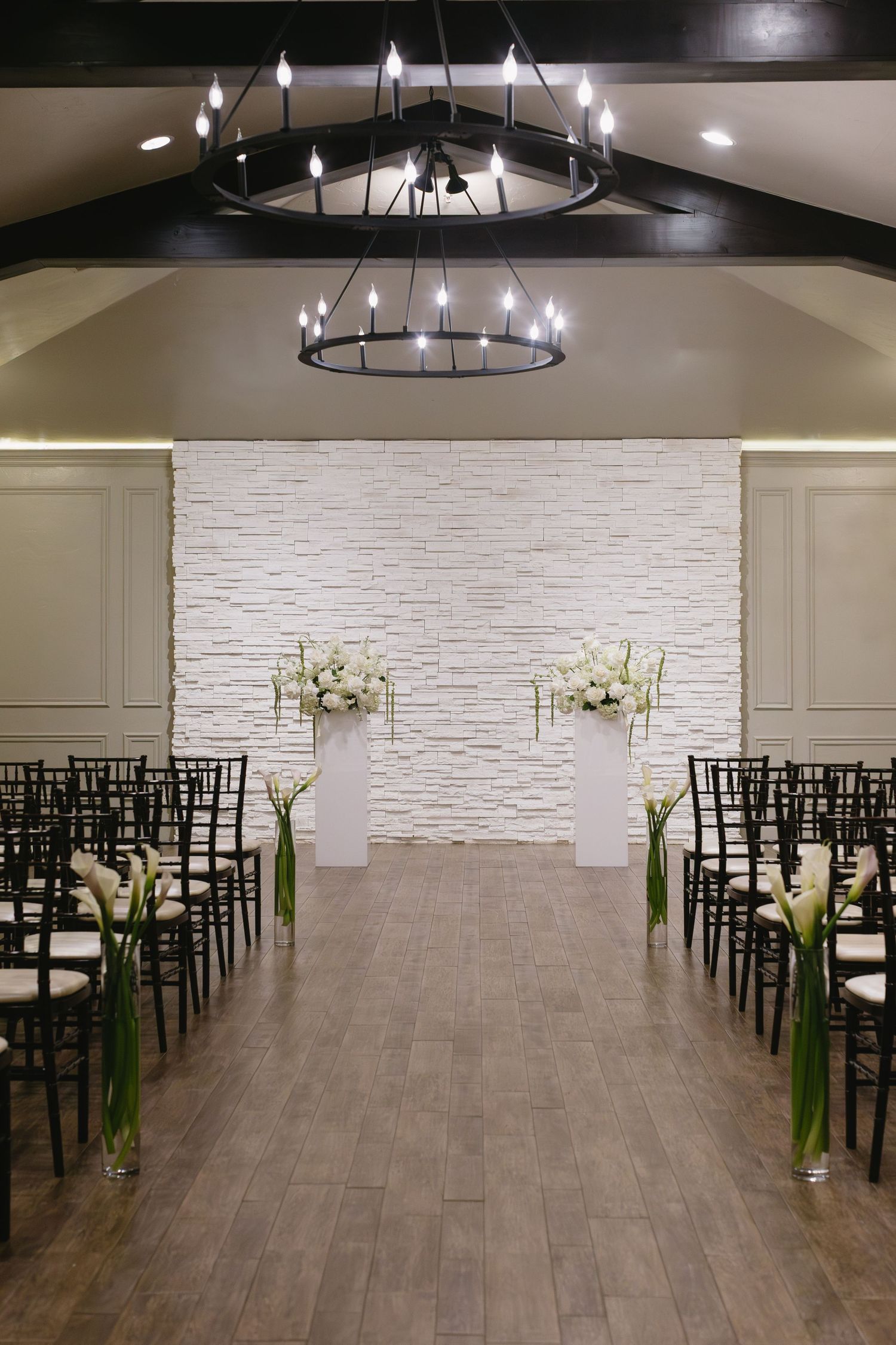 Wedding ceremony aisle with black chairs, circular chandeliers, and white floral arrangements on pedestals.