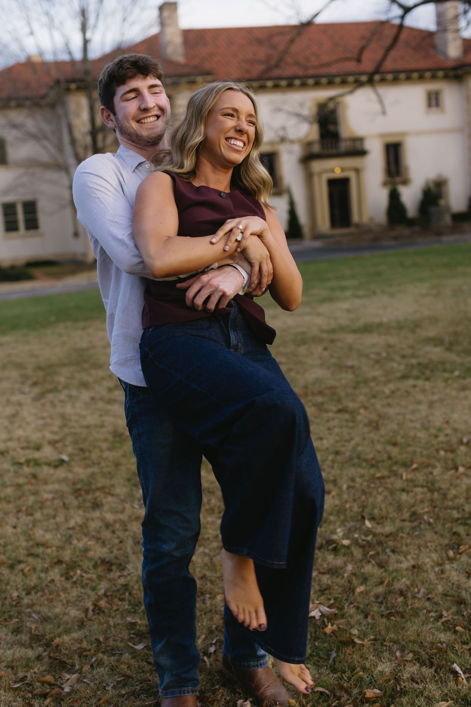 A joyful couple embraces and laughs together on a lawn near historic architecture.