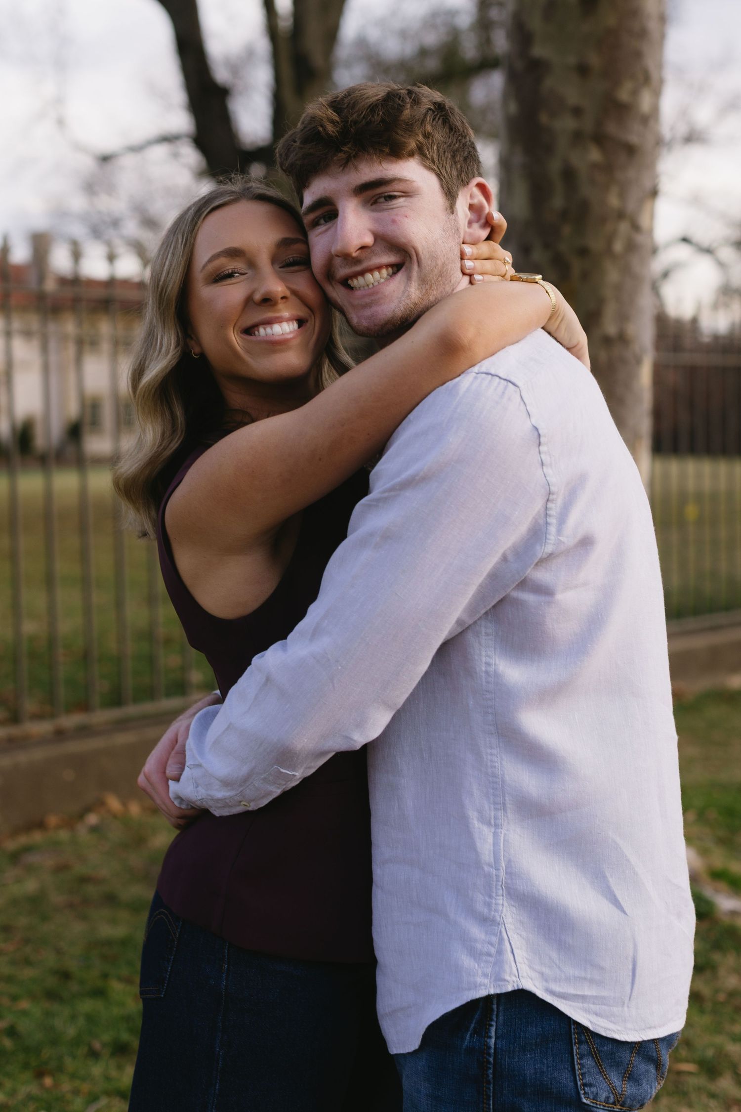 A couple embraces and smiles outdoors with trees and architecture in the background.