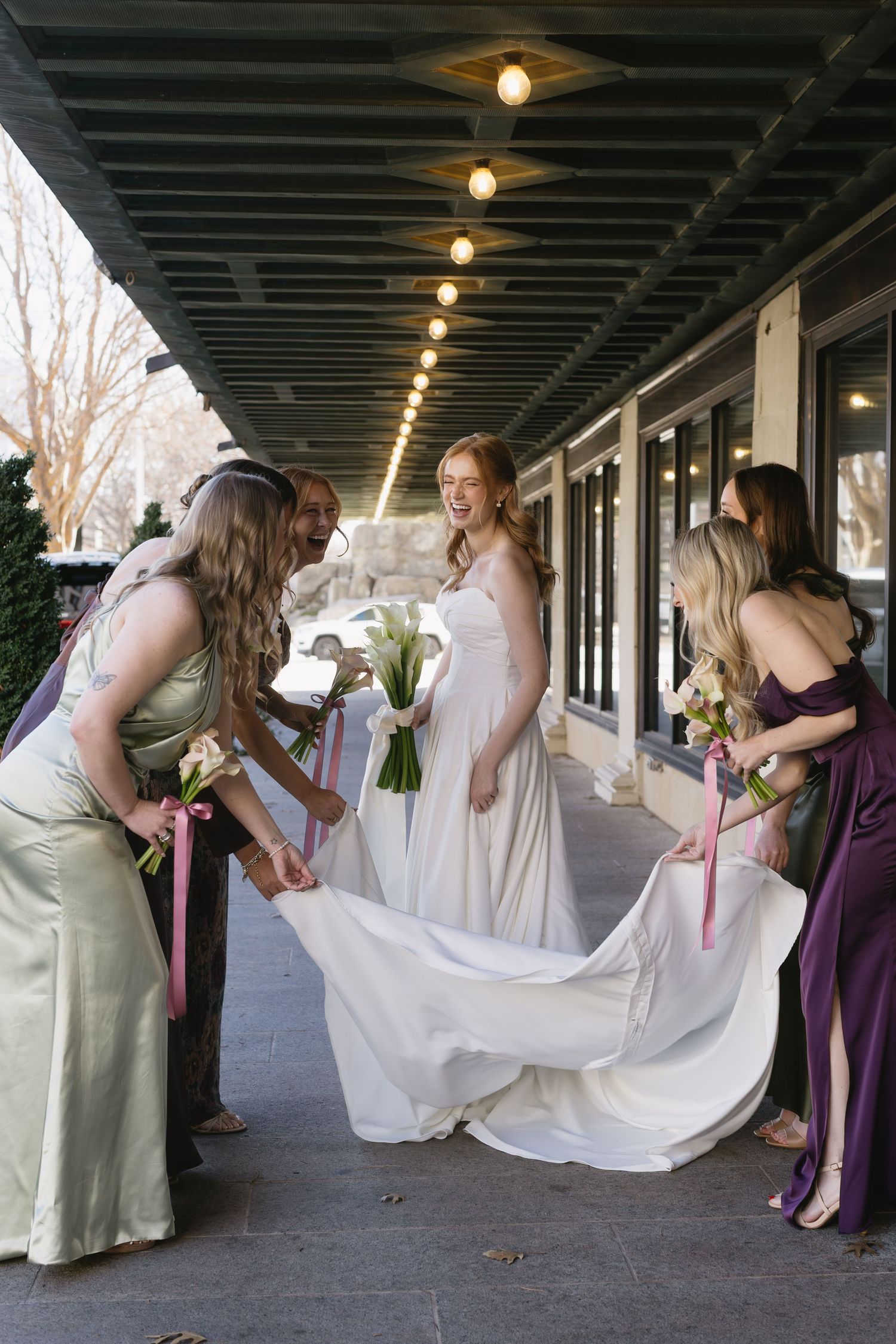 Bridesmaids in purple and sage dresses help adjust the bride's gown train outdoors.
