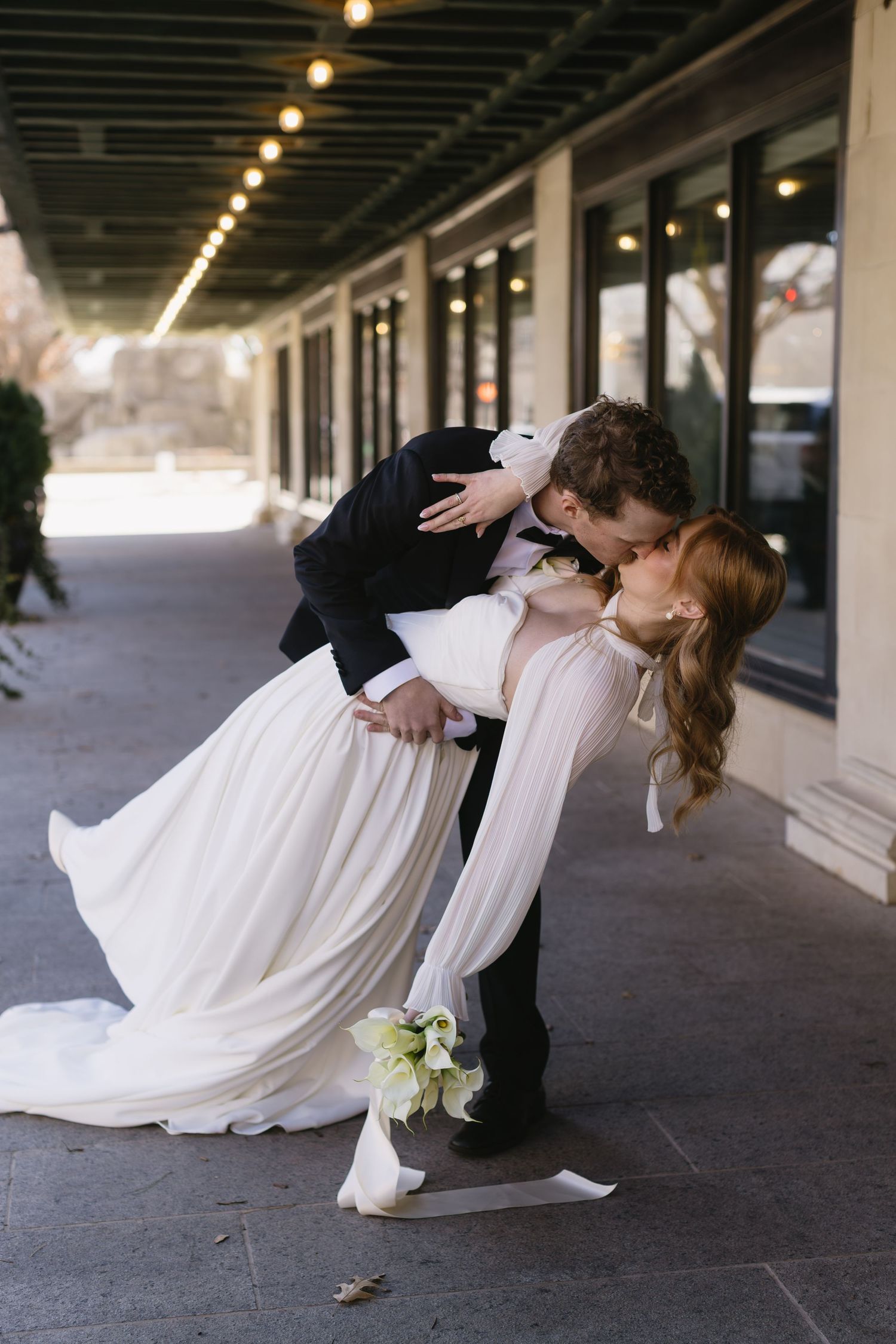 A romantic dip pose between newlyweds in formal attire under a covered walkway with string lights.
