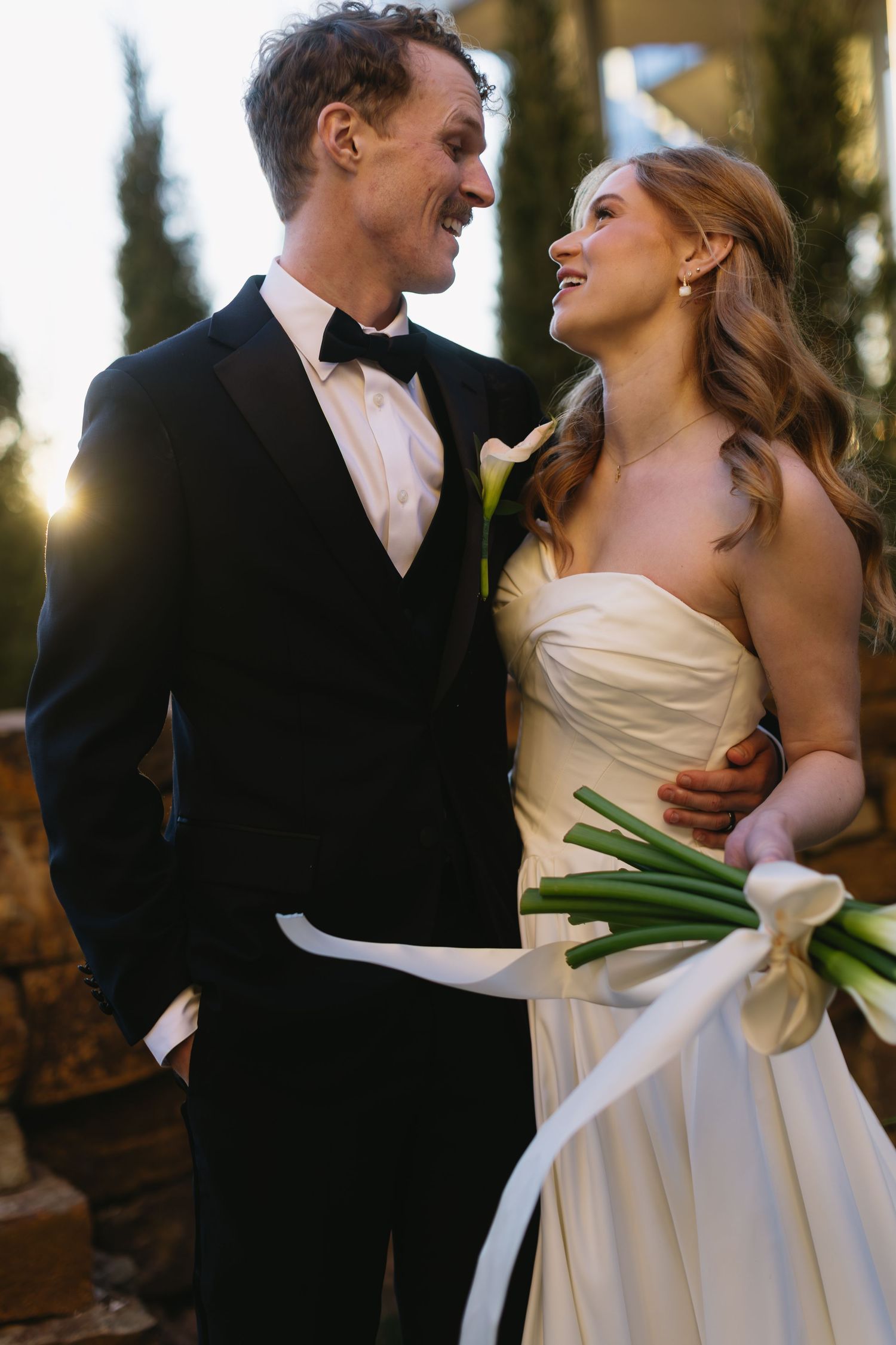 A couple shares a romantic laugh while holding a bouquet of white calla lilies at sunset.
