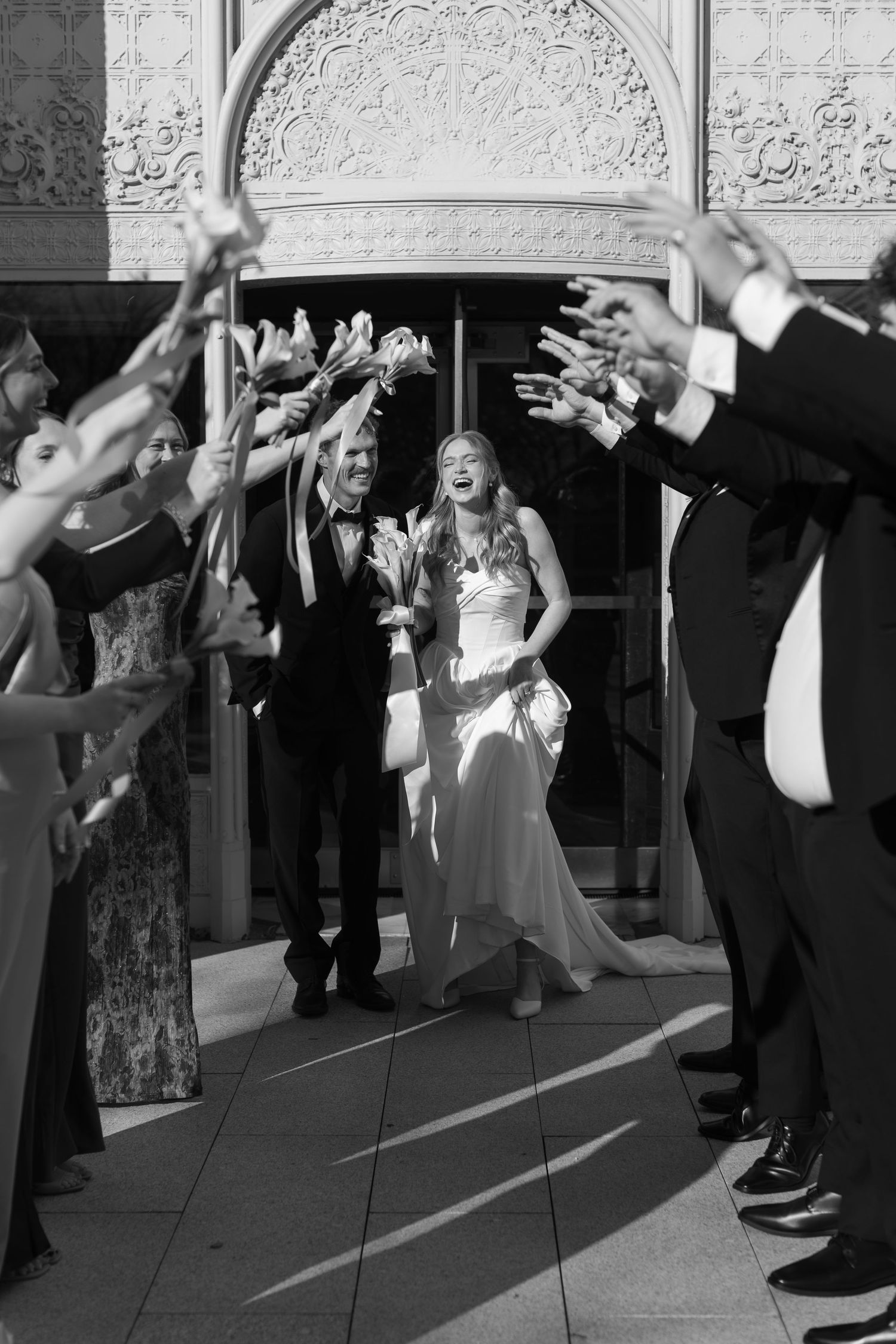 Wedding guests and bridal party celebrate with raised hands in front of ornate architectural backdrop.