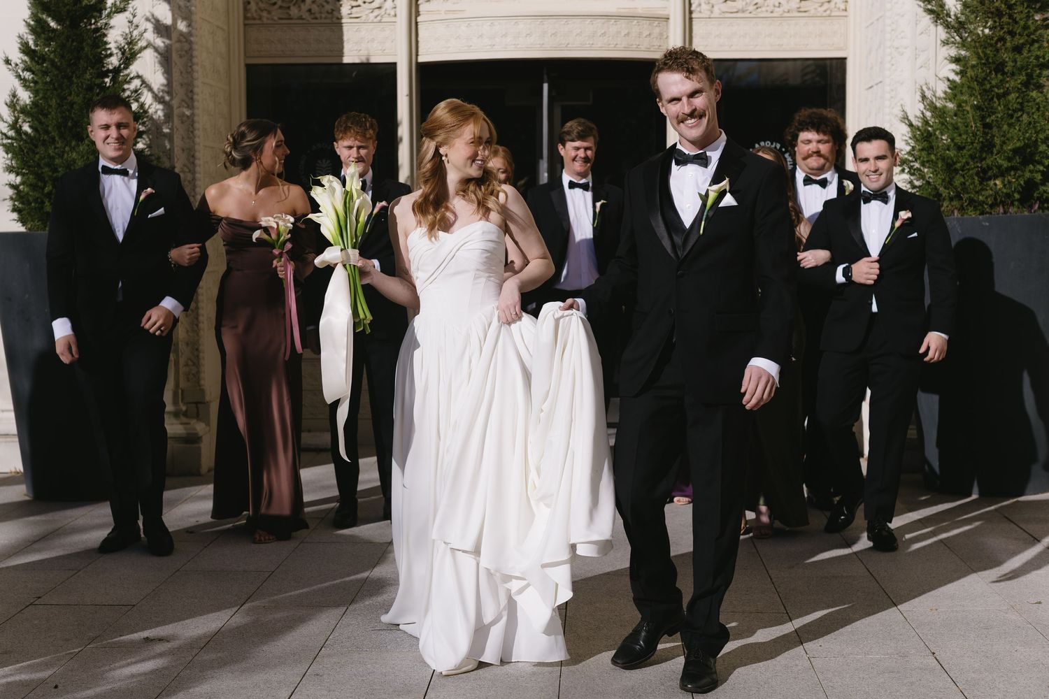 Wedding processional with groomsmen in black tuxedos outside an ornate building.