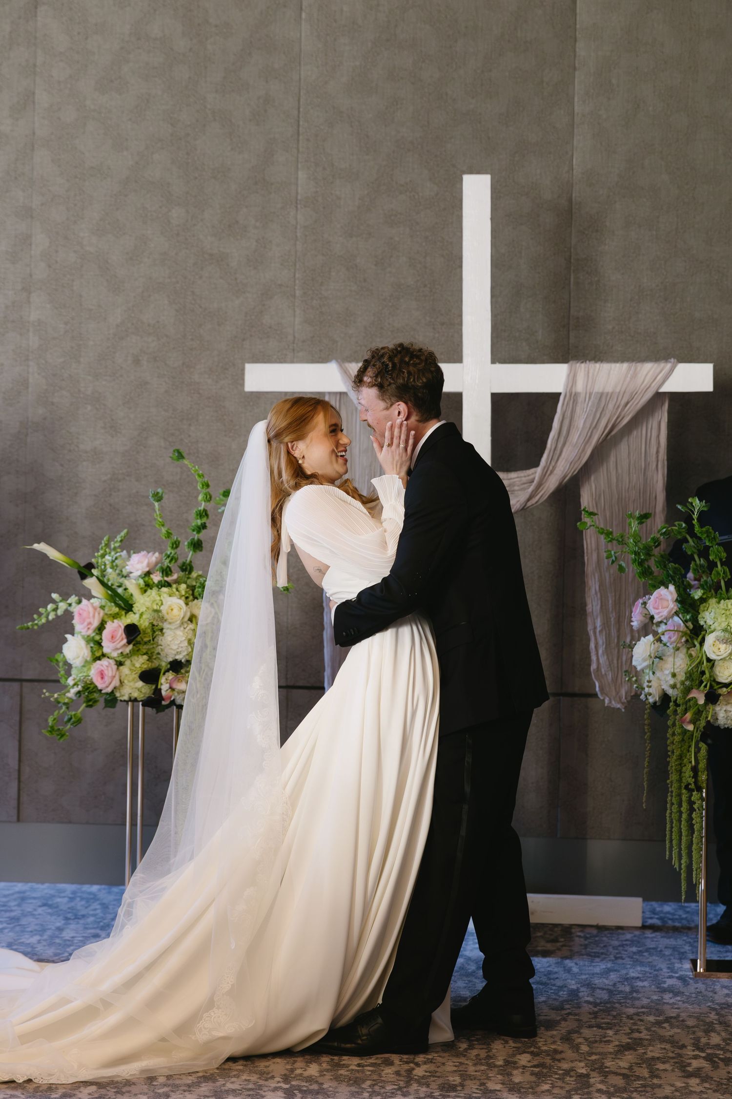 A wedding couple shares an intimate moment near floral arrangements and a white cross.