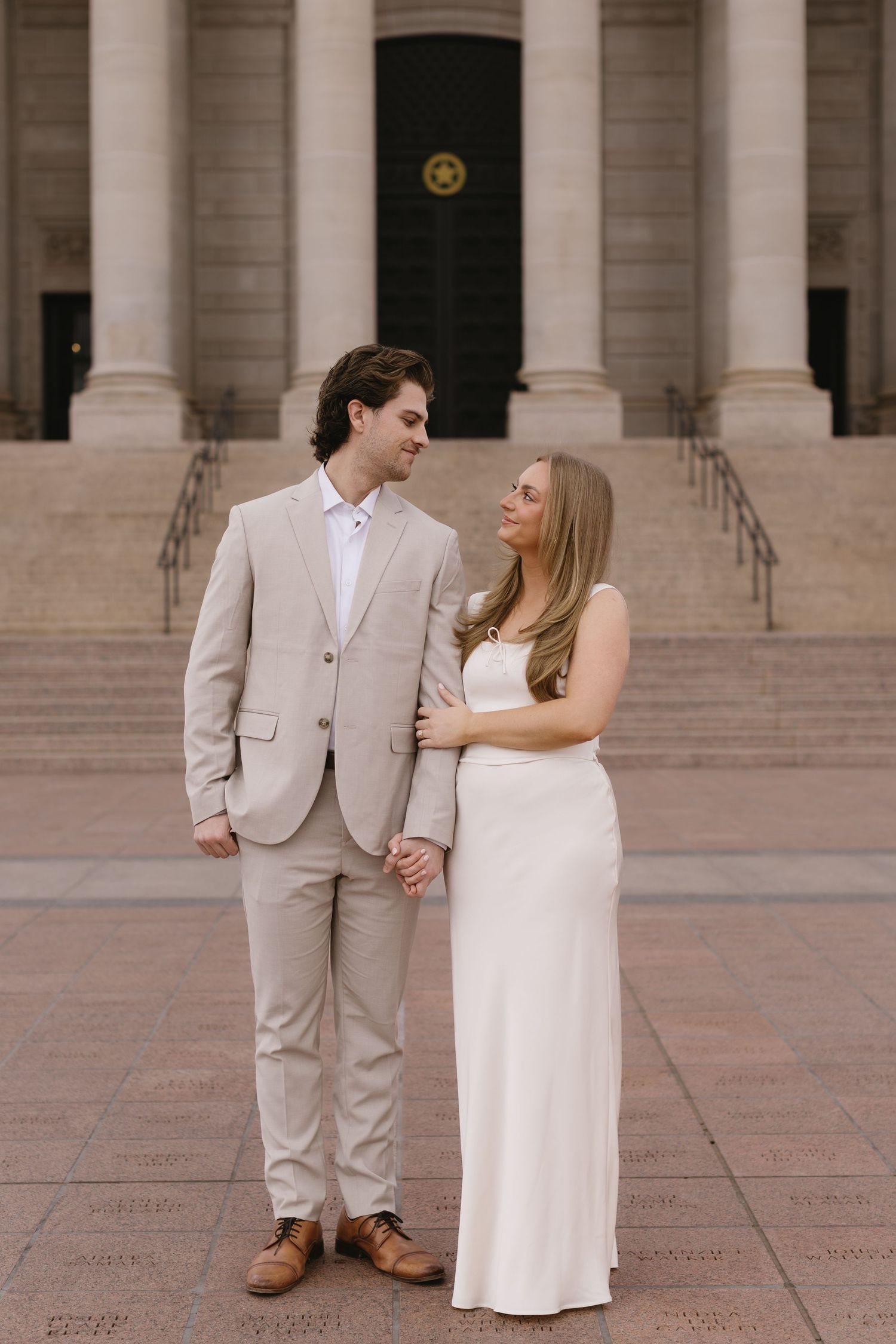 A couple in coordinated beige and white outfits poses together on a grand stone plaza.