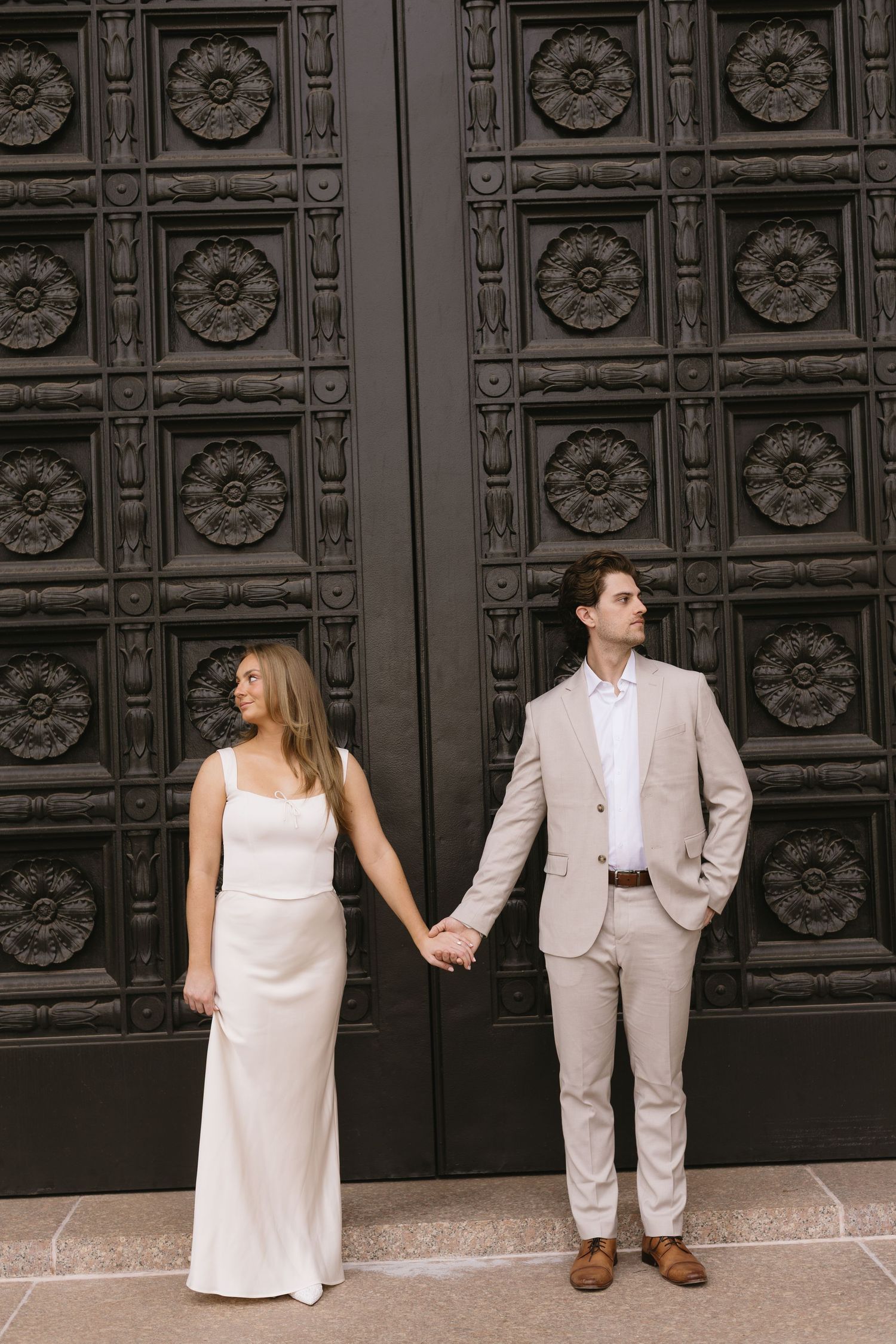 A couple in coordinated beige and white formal wear poses against an ornate dark door with floral patterns.