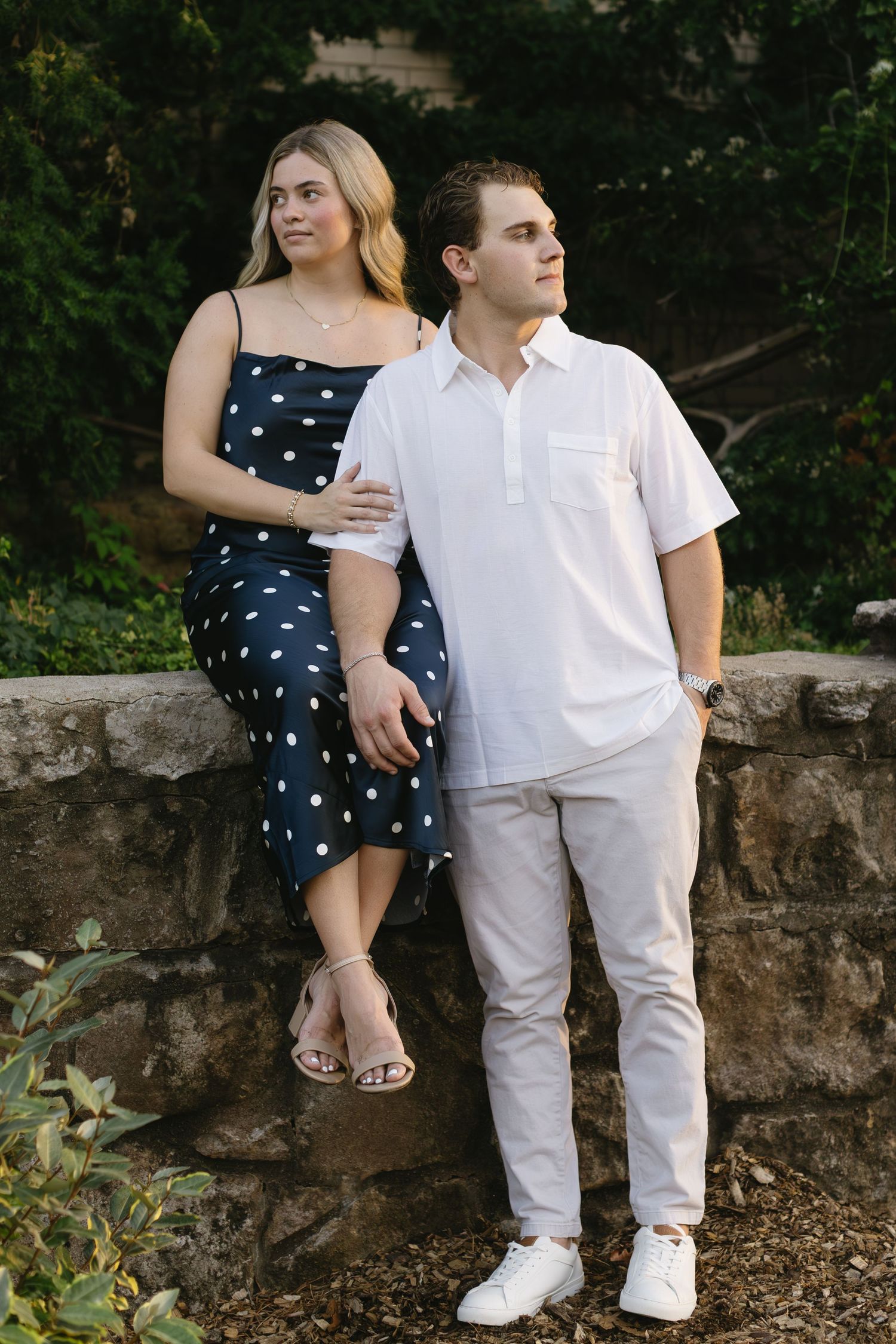 A couple sits together on a stone wall, wearing a navy polka dot dress and white casual outfit.